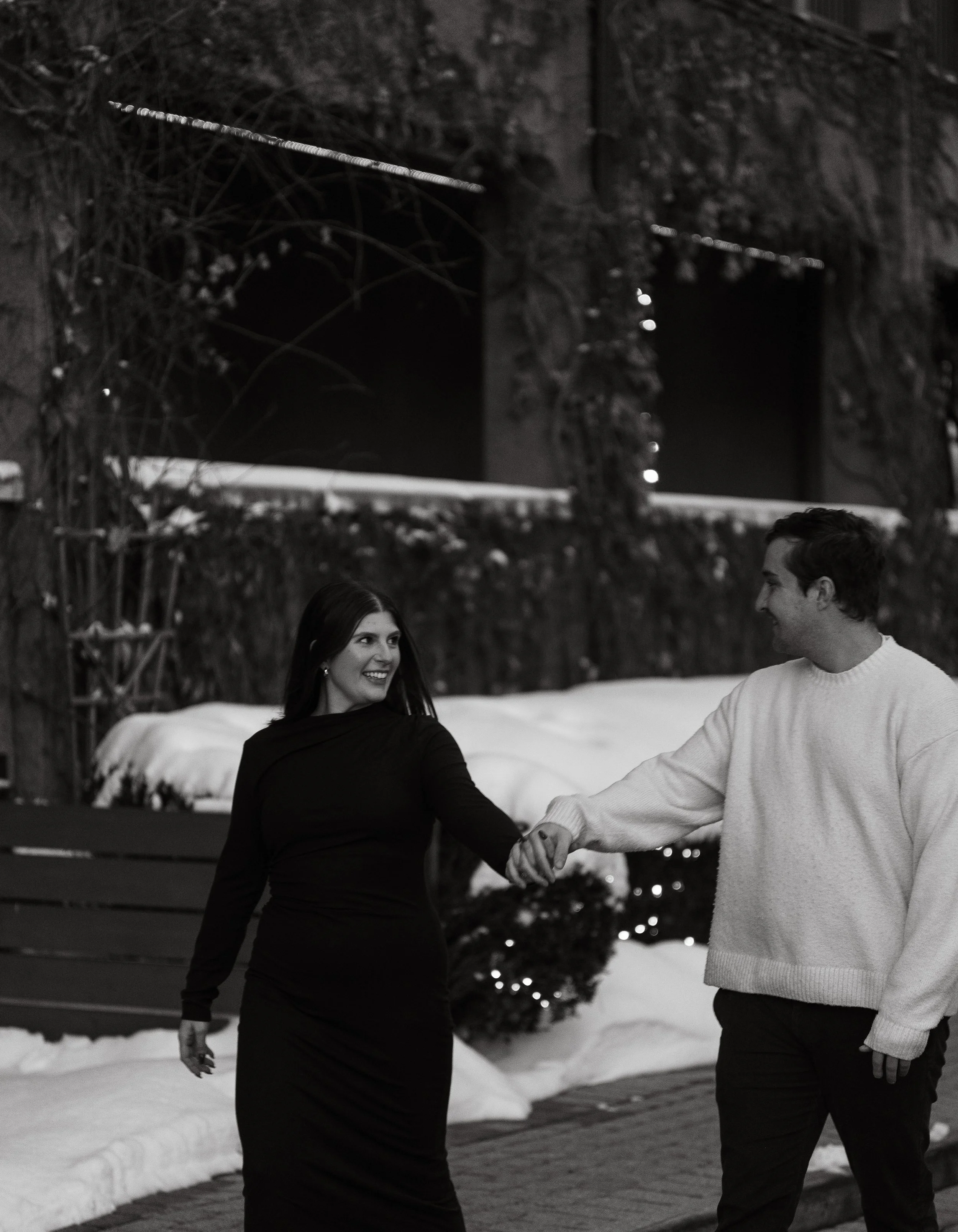 A woman in a black dress and a man in a light sweater are holding hands and smiling at each other outdoors at night, with snow on the ground and holiday lights in the background.