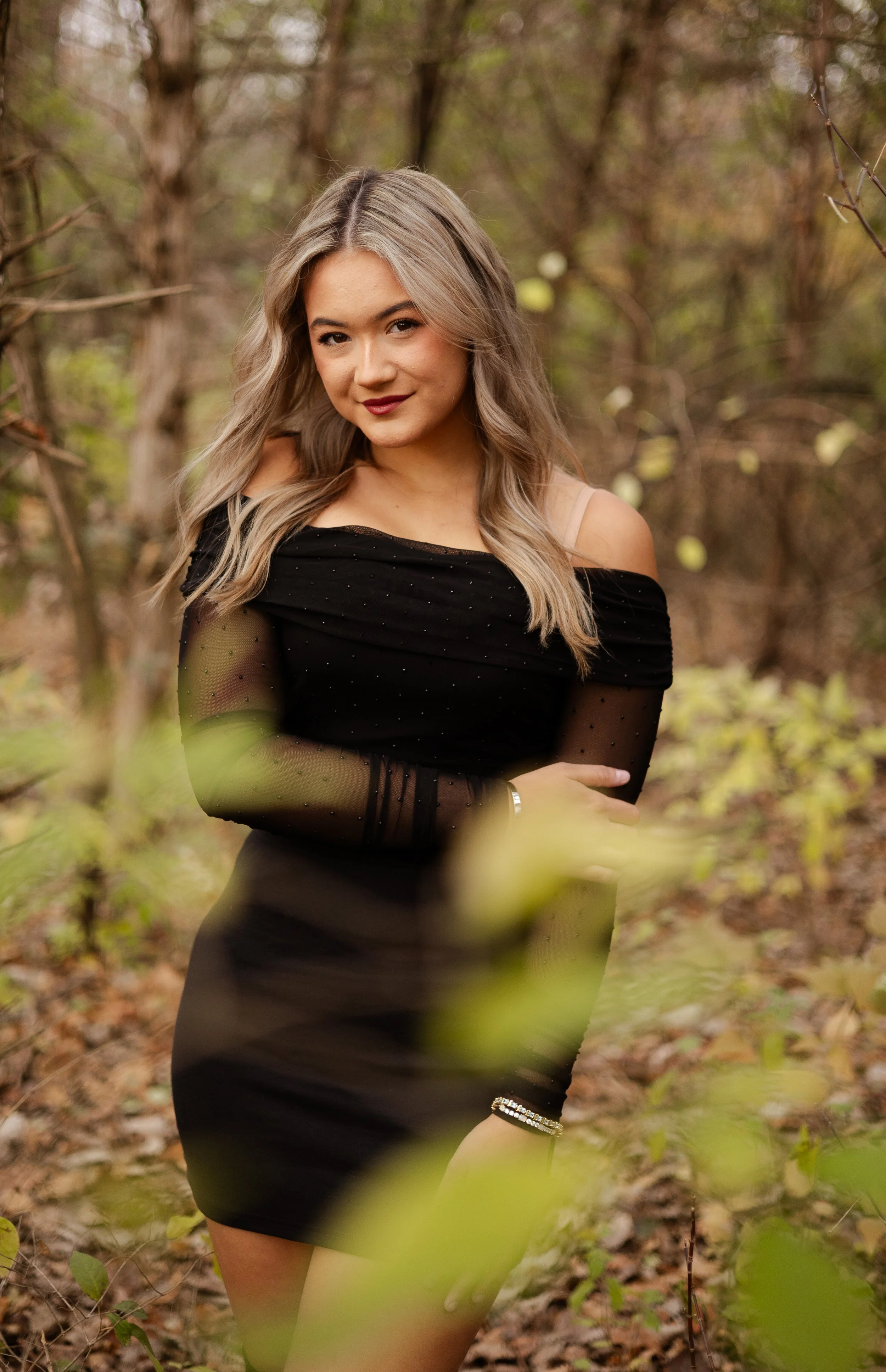 A young woman with long blonde hair and light makeup, wearing a black off-shoulder dress with sheer long sleeves and a bracelet, stands outdoors in a forested area with trees and leaves in the background.