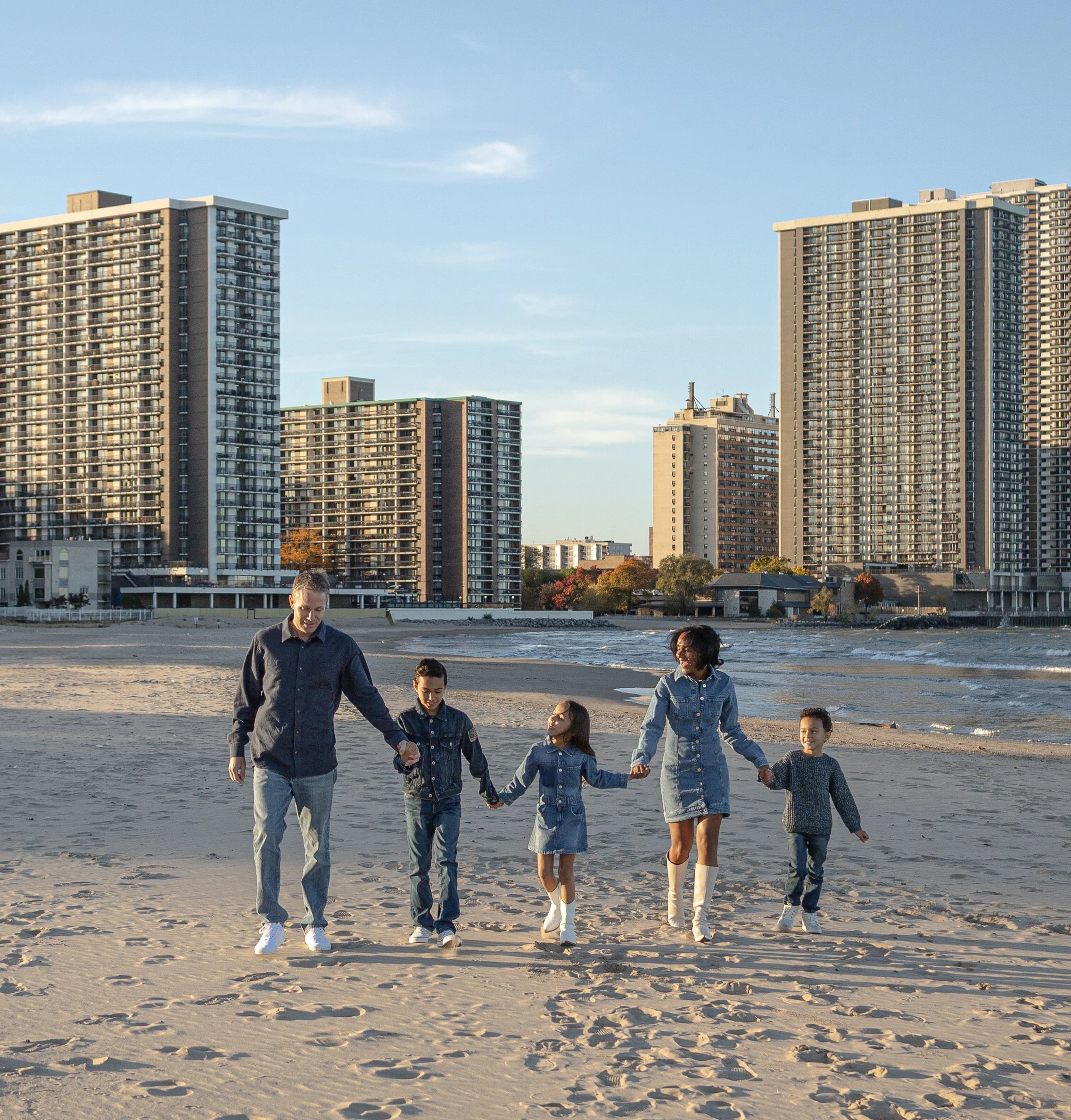 A family walks on the beach holding hands with high-rise buildings in the background and a partly cloudy sky.