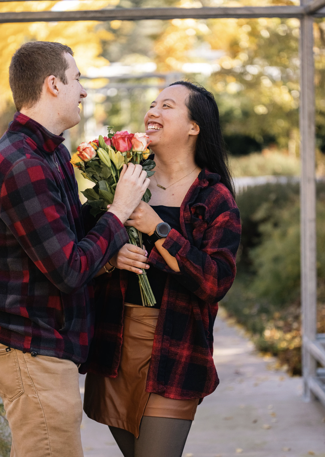 A man is giving a woman a bouquet of flowers outdoors during fall, with trees with yellow leaves in the background. They are smiling at each other.