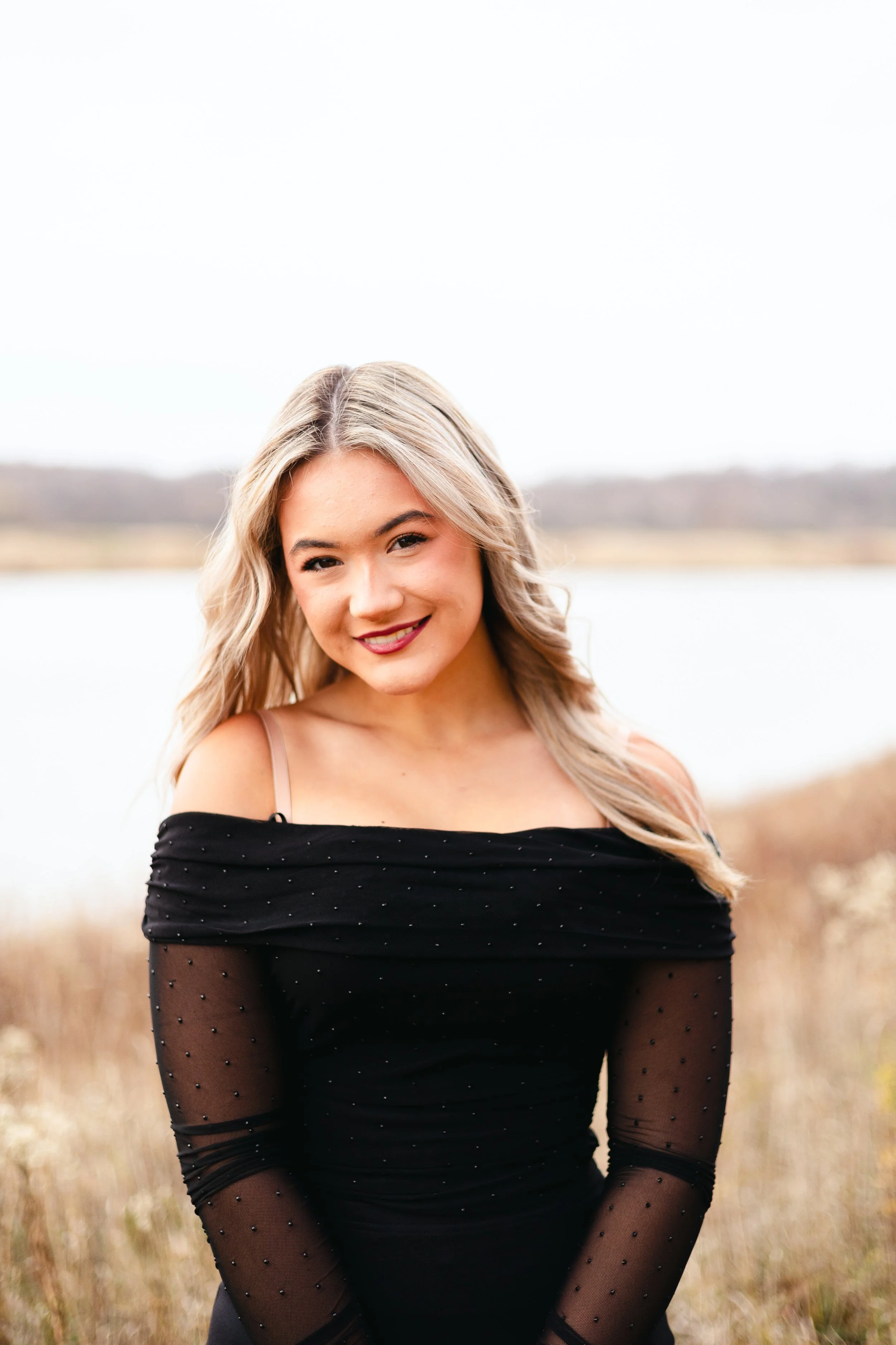 A young woman with blonde hair wearing a black off-shoulder top with sheer, dotted sleeves standing outdoors near a body of water and grass.