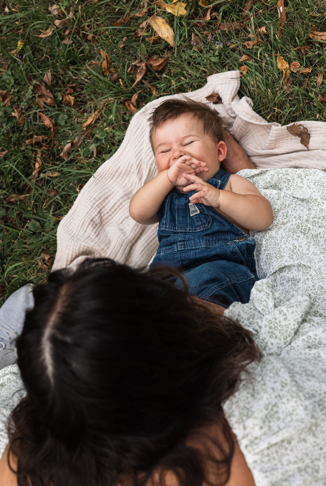 A young child lying on a blanket outdoors, laughing and covering his nose with his hand, surrounded by grass and fallen leaves.