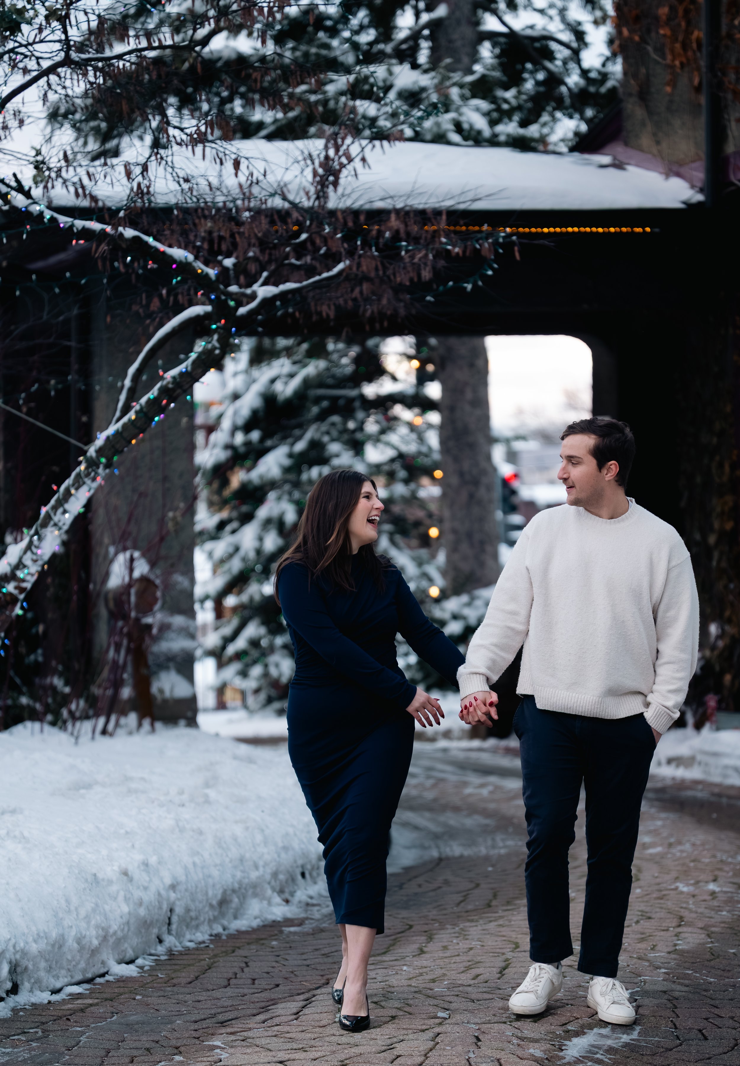 A couple dressed in cold-weather clothing walking hand-in-hand on a snowy sidewalk, smiling and looking at each other.