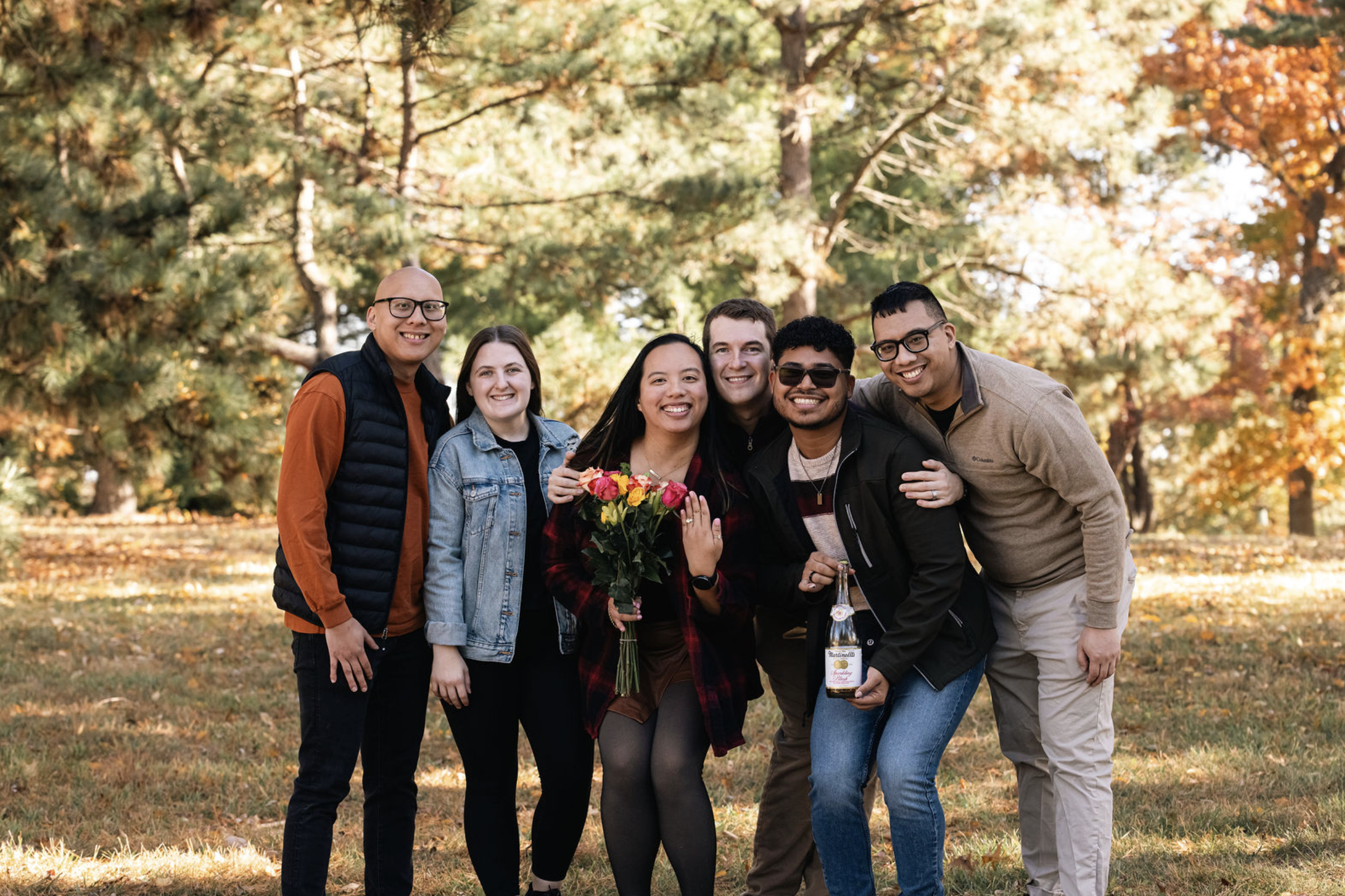 Group of six friends smiling and posing together outdoors in a park during fall, with trees and autumn foliage in the background. One person in the center holds a bouquet of flowers, and another holds a bottle of alcohol.