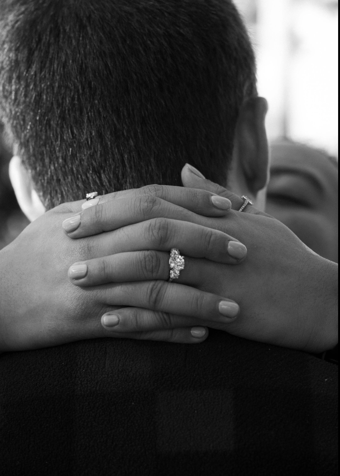 A couple embraces, with one person holding the other from behind, showing their hands with rings and a wedding band, black and white photo.