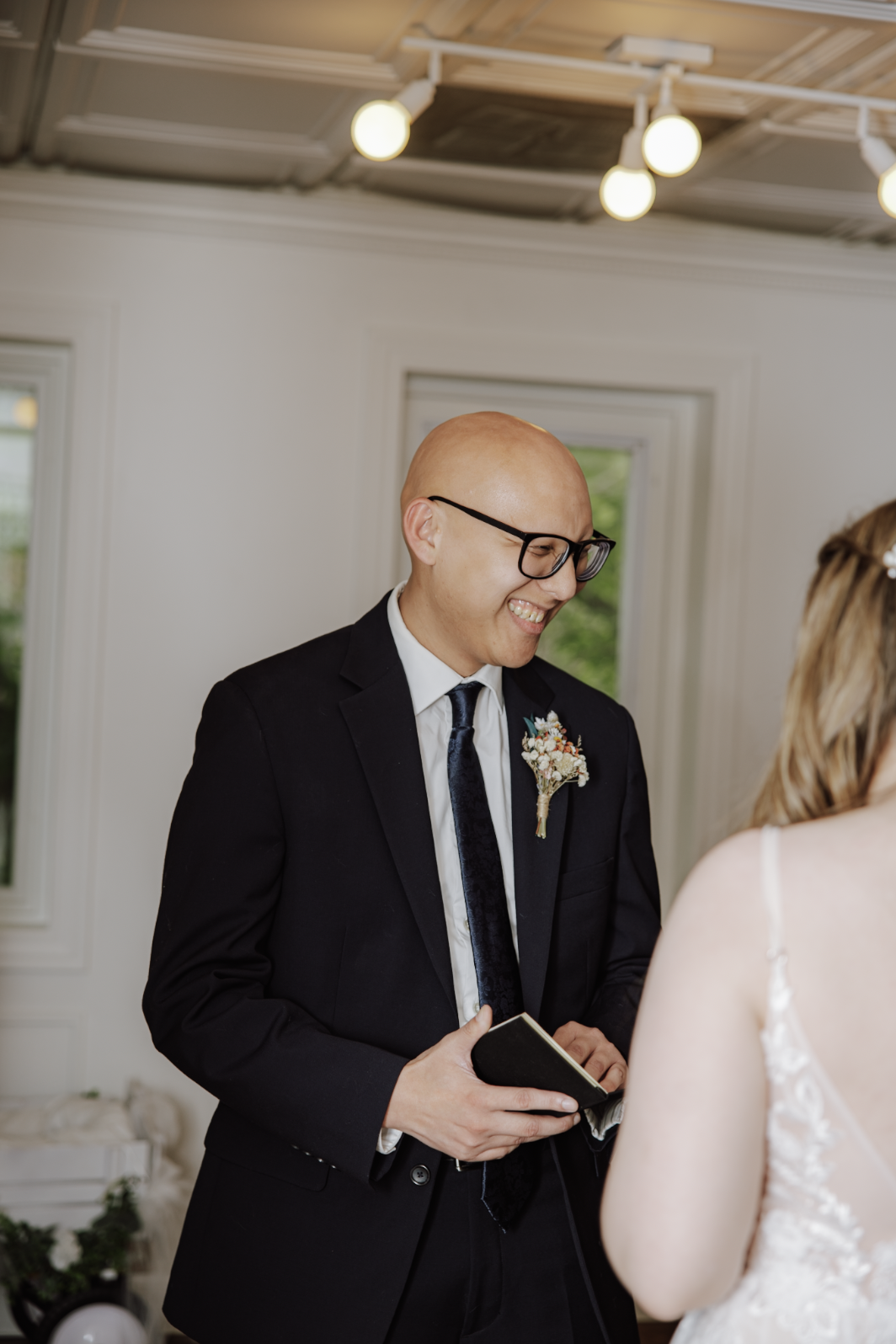 A man wearing glasses and a black suit with a white shirt and tie, holding a small book, smiling during a wedding ceremony, standing in a room with white walls and large windows.