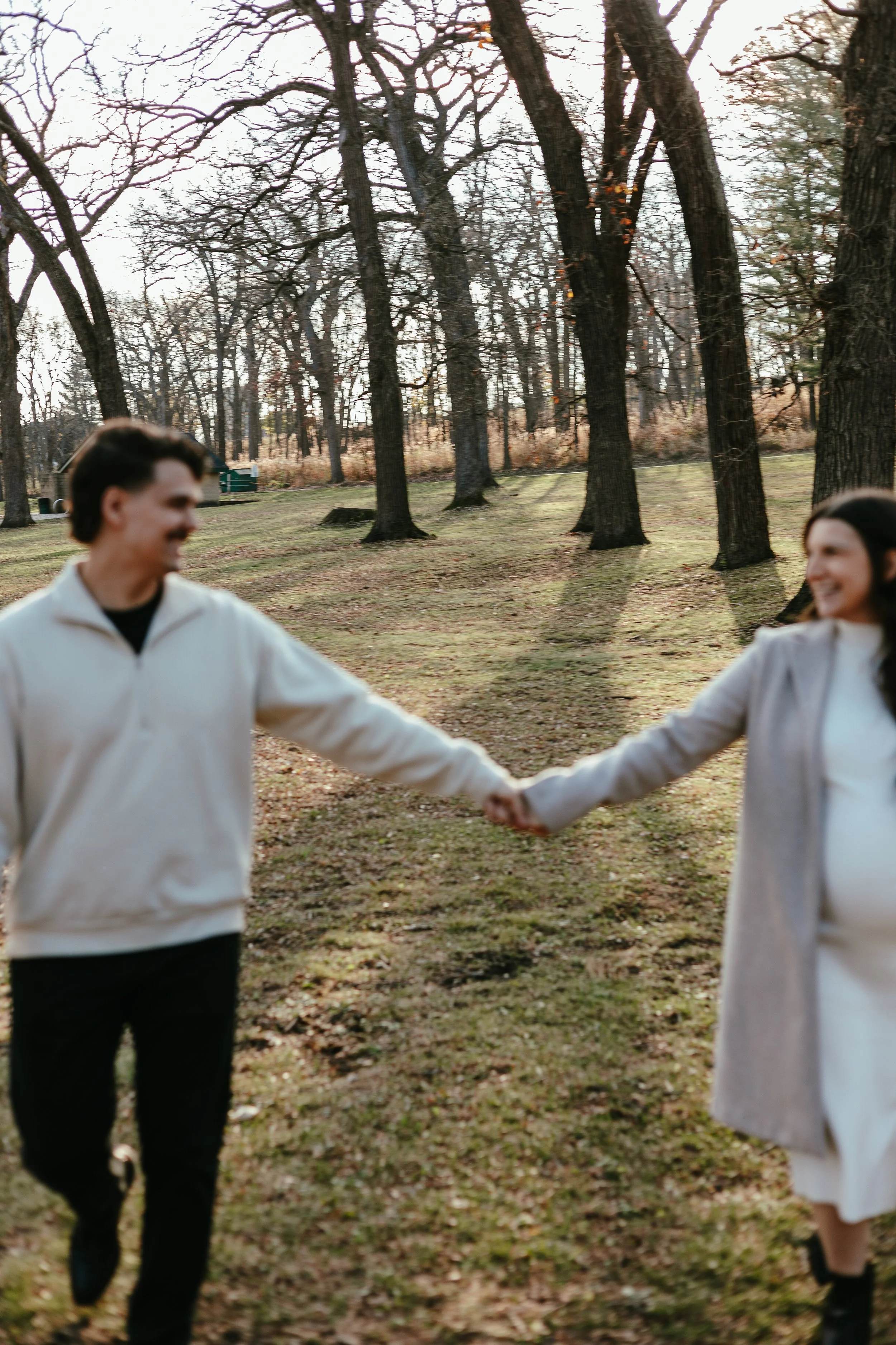 A man and woman holding hands and smiling at each other outdoors in a park with leafless trees.