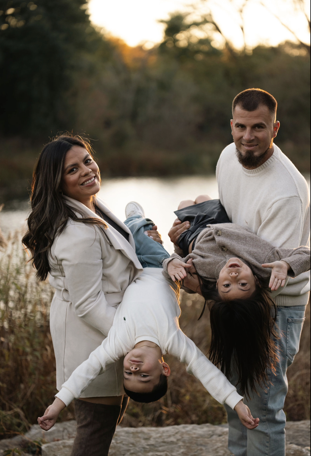 A family of four enjoying outdoors near a lake at sunset. The mother holds a girl upside down, the father holds a boy, and the family smiles happily.