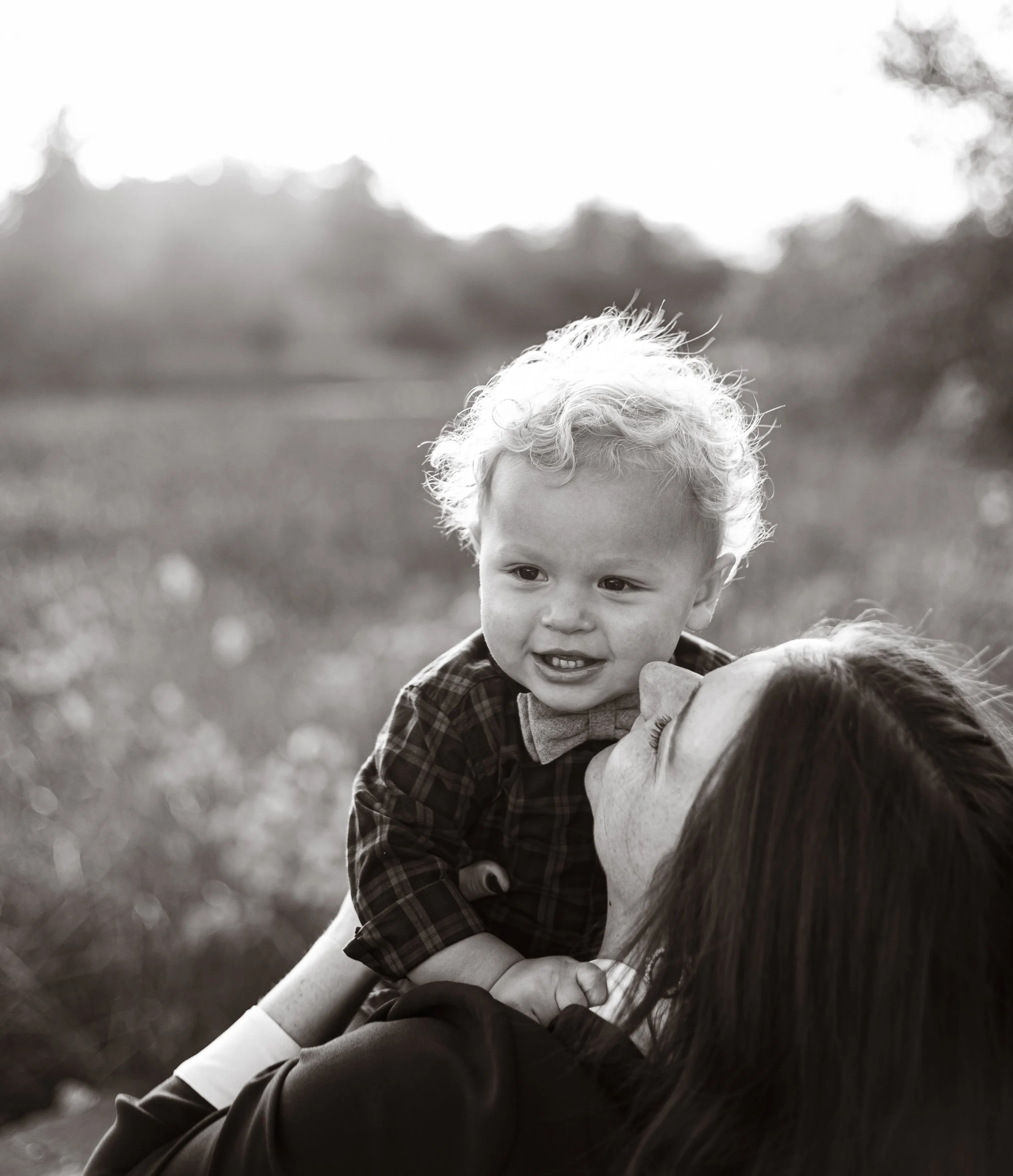 A young boy smiling outdoors, being embraced by an adult, with blurred trees and grass in the background.