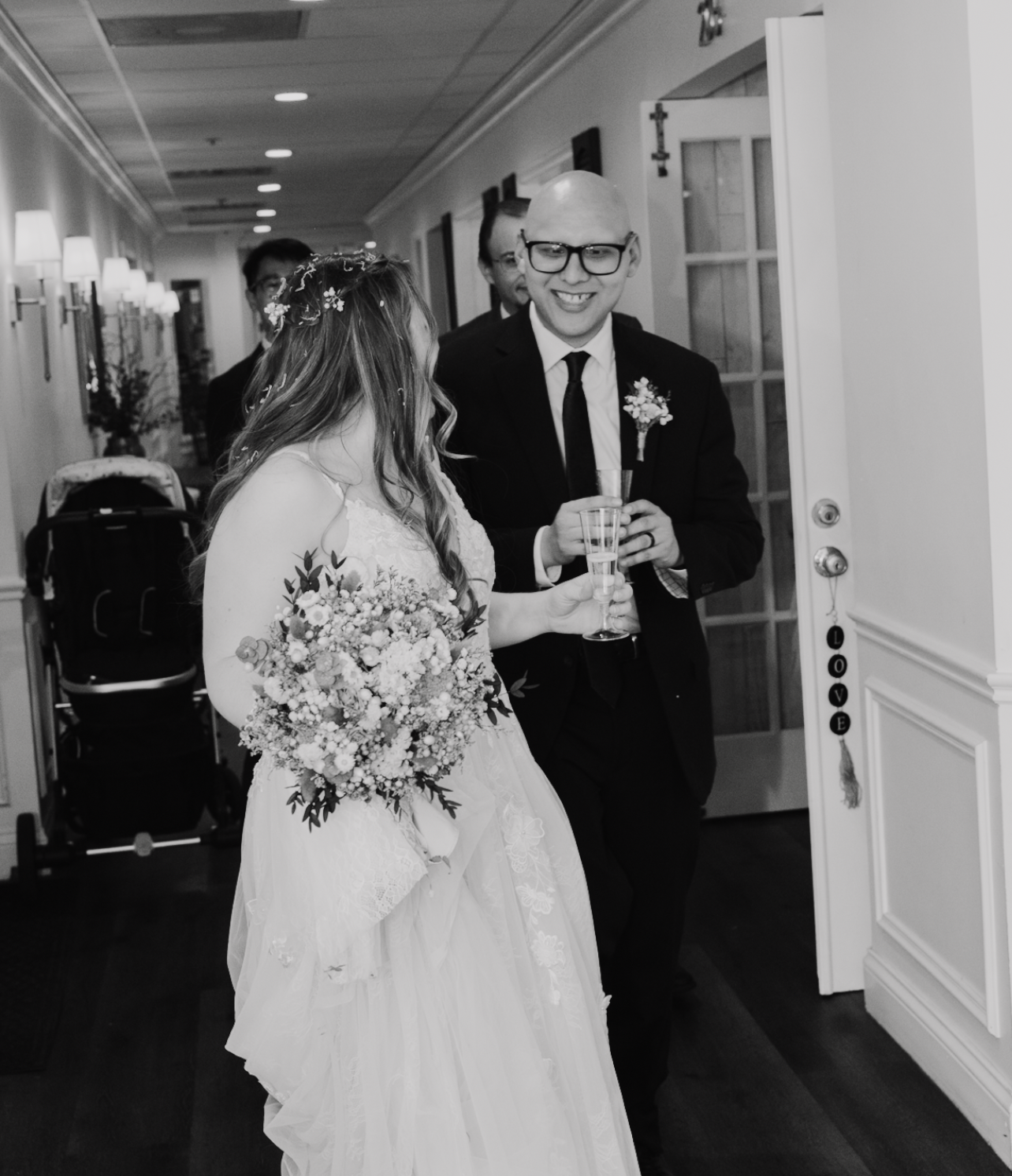 A bride and groom smiling and holding glasses at a wedding reception. The bride is wearing a gown and holding a bouquet, while the groom is dressed in a suit with a boutonniere. There are guests in the background and a door decorated with a 'LOVE' si