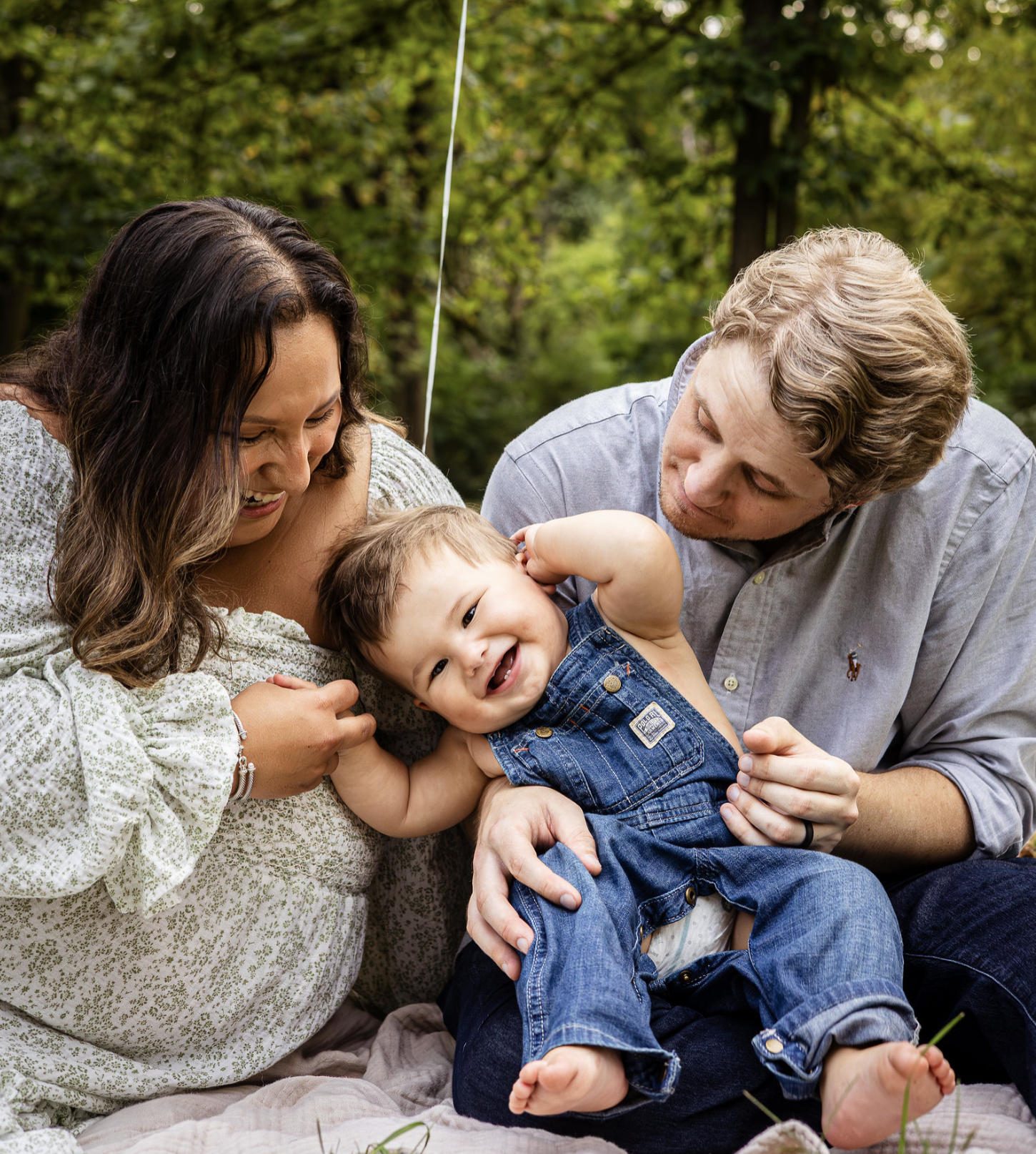 A family of three, an Asian woman, a white man, and a smiling young boy, lying on a blanket outdoors with trees in the background. The boy is playing with the woman, and both adults are smiling and looking at him.
