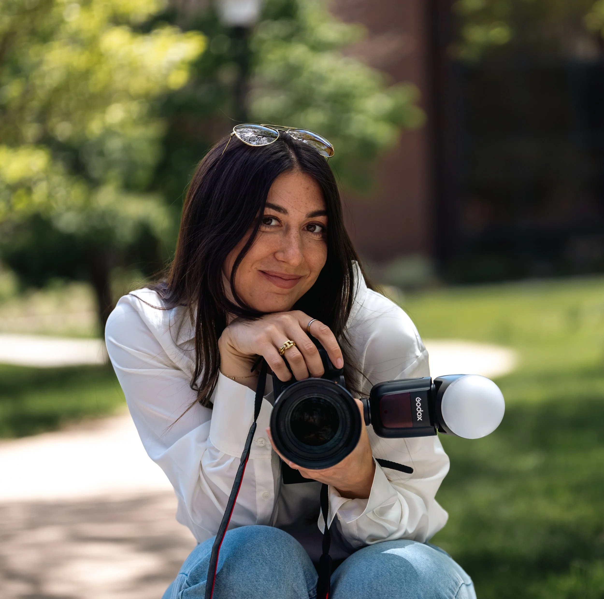 A young woman with long dark hair and freckles, wearing a white jacket and blue jeans, is sitting outdoors on a park bench. She is holding a camera with a large lens on her lap, and sunglasses are resting on her head. The background is blurred greenery and sunlight.