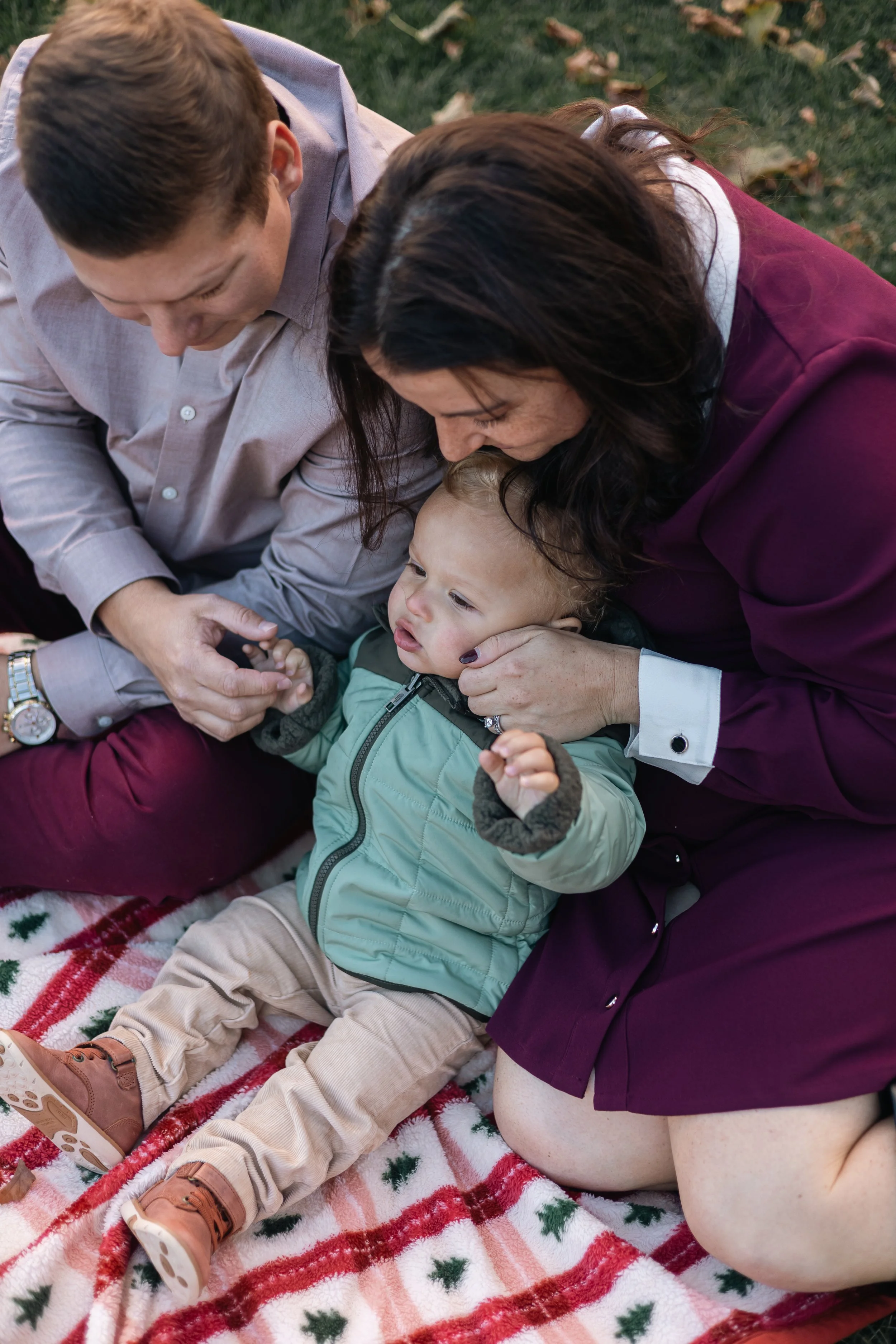 Family sitting on a Christmas-themed blanket outdoors, holding hands and cuddling, with fallen leaves on the grass in the background.