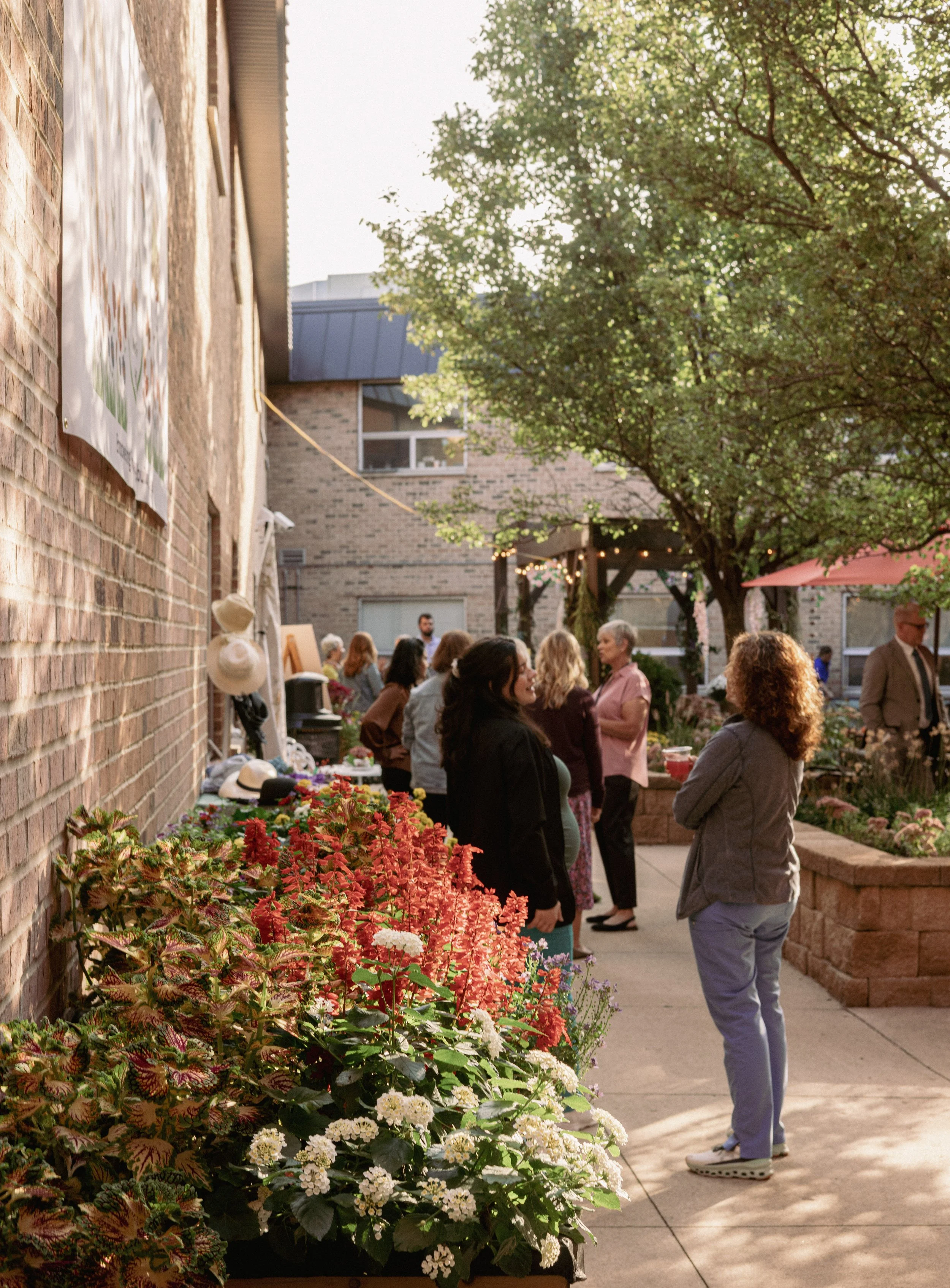 People gathering outdoors in a courtyard with flowers, trees, and string lights, engaging in conversation during late afternoon or early evening.