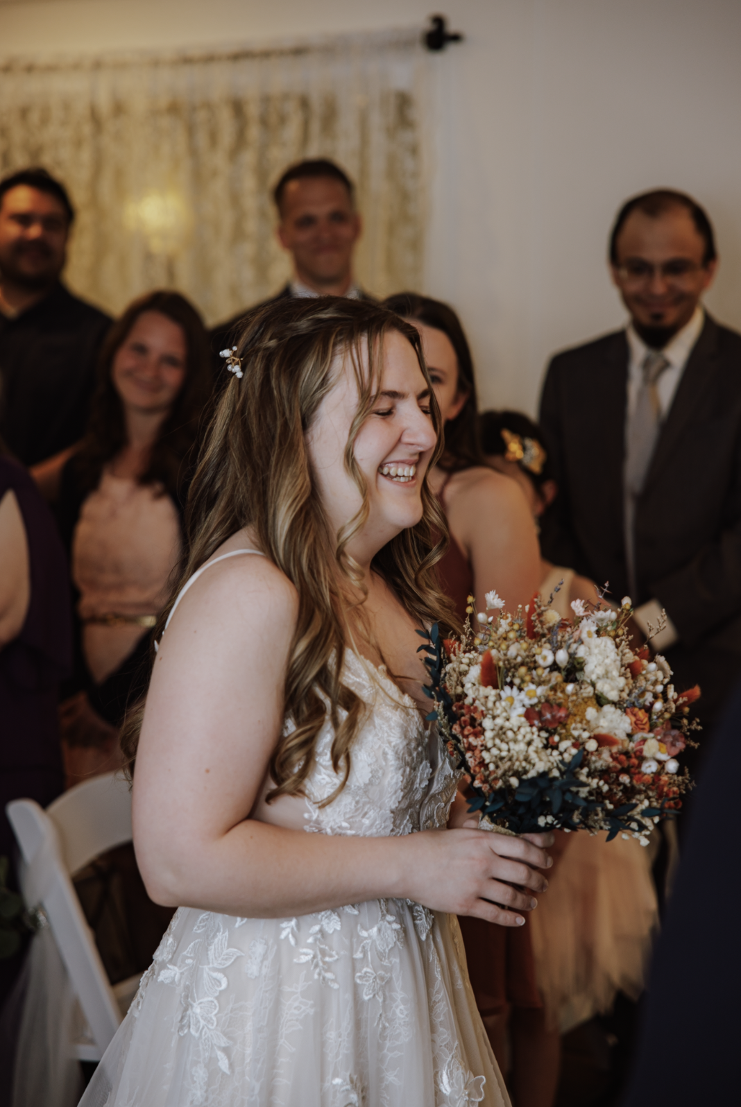 A bride with long wavy hair holding a bouquet of flowers, smiling at her wedding surrounded by friends and family.