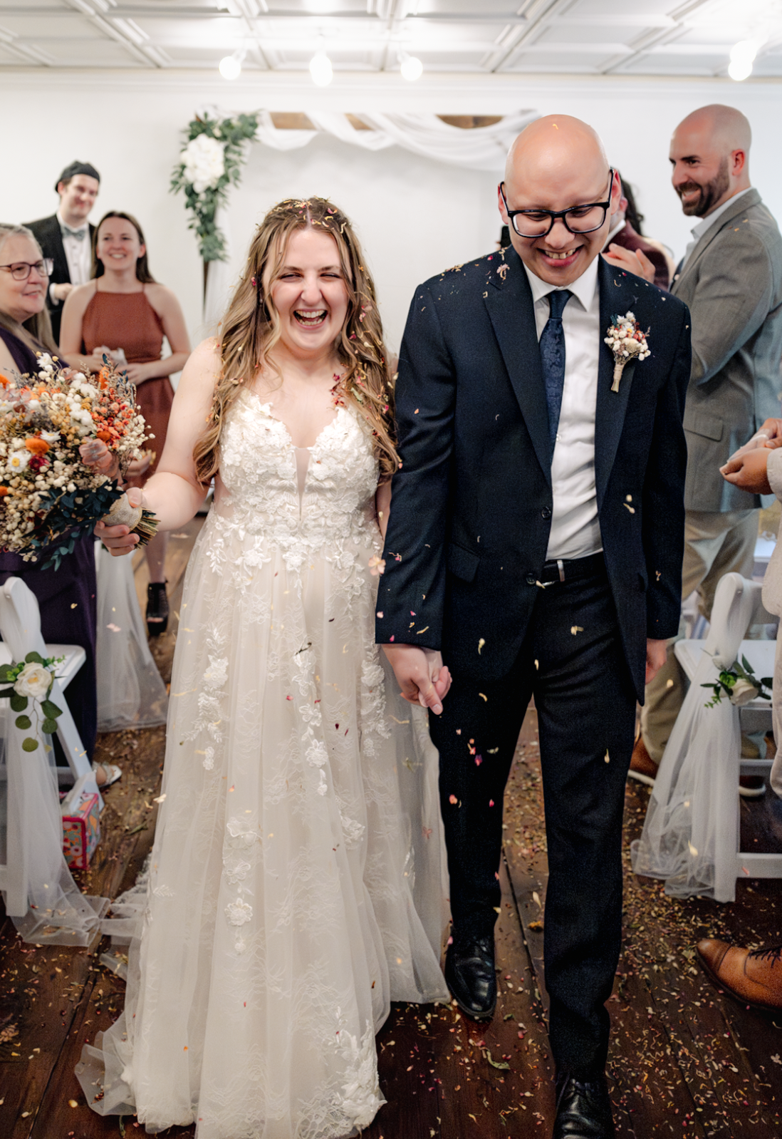 A bride and groom smiling and walking hand in hand at a wedding ceremony, surrounded by guests, with confetti falling around them.