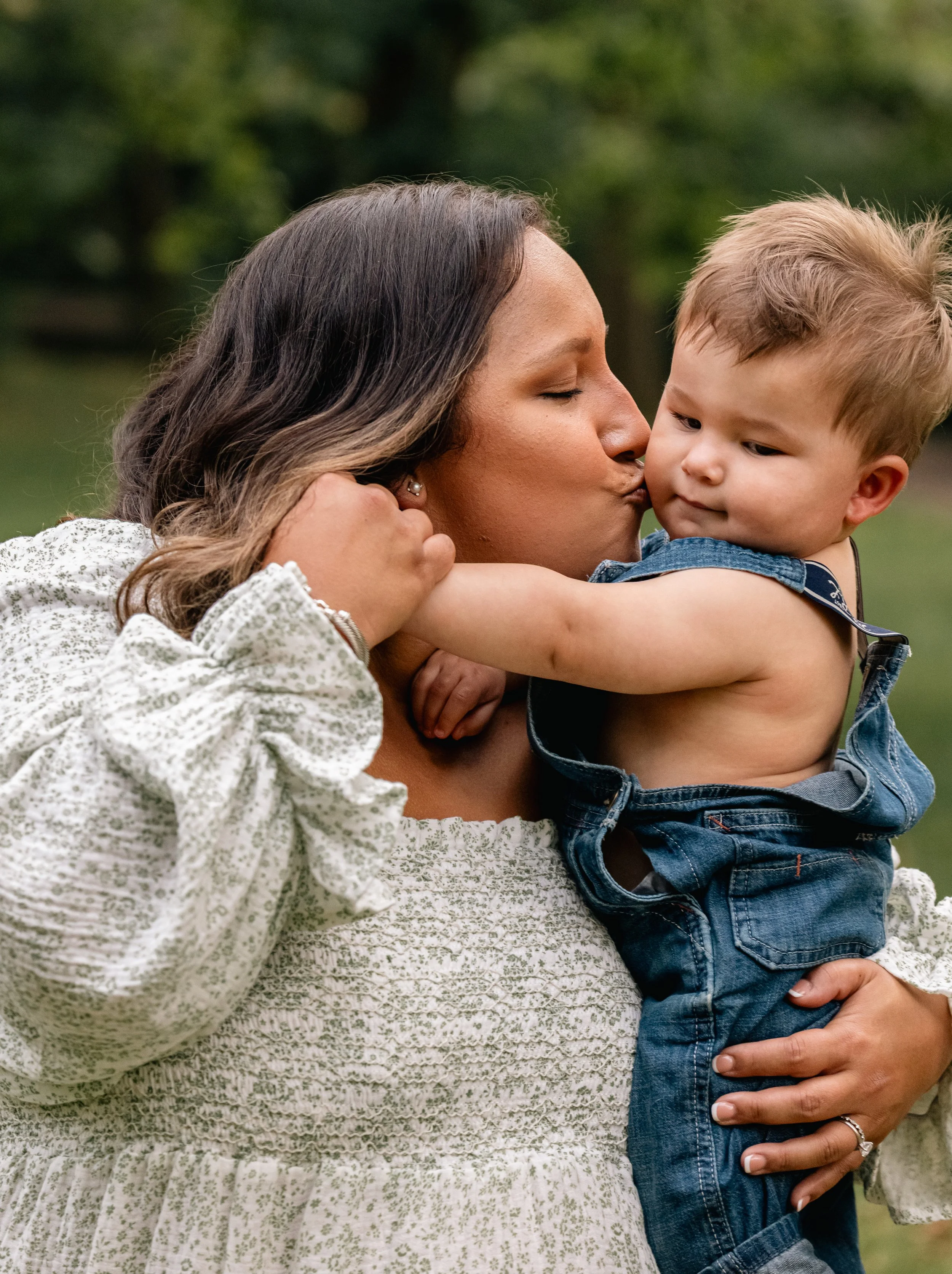 A woman with closed eyes, holding a young boy and kissing his cheek outdoors. The woman has dark hair and is wearing a white patterned blouse. The boy has short, light hair and is wearing a denim vest and jeans.