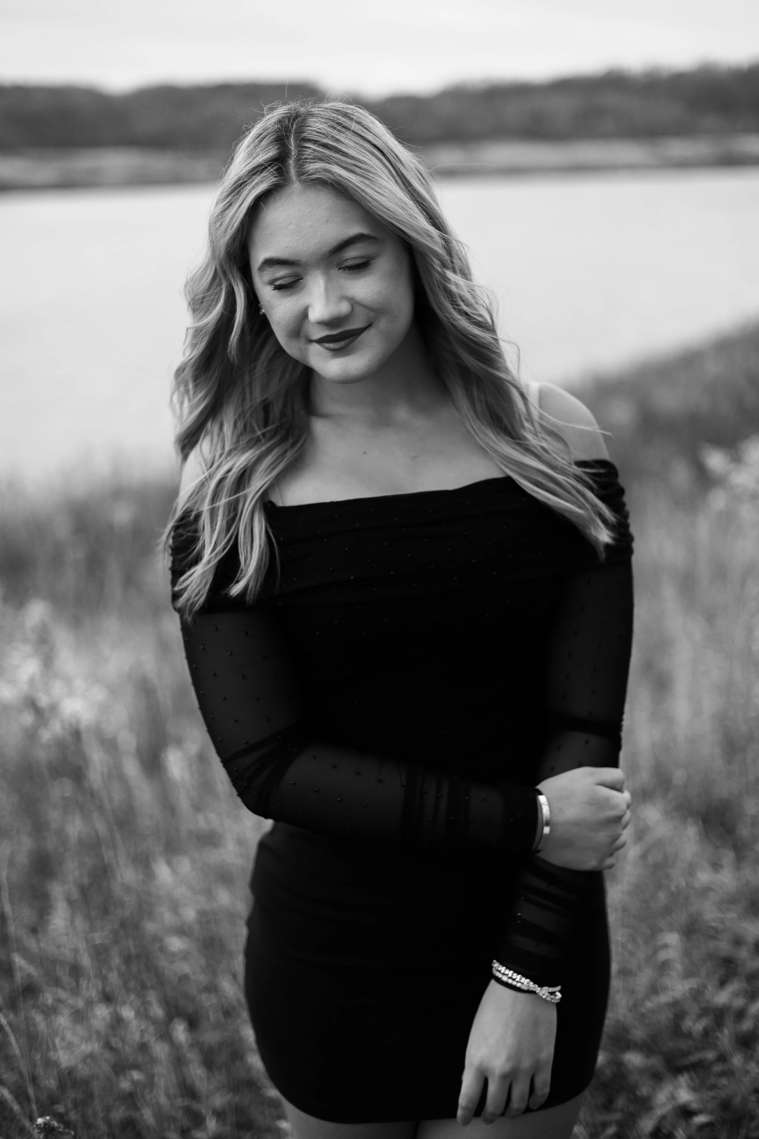 A black and white photo of a young woman with long wavy hair, smiling slightly with her eyes closed, outdoors near a body of water and grassy area in the background, wearing a dark off-the-shoulder top and a bracelet.