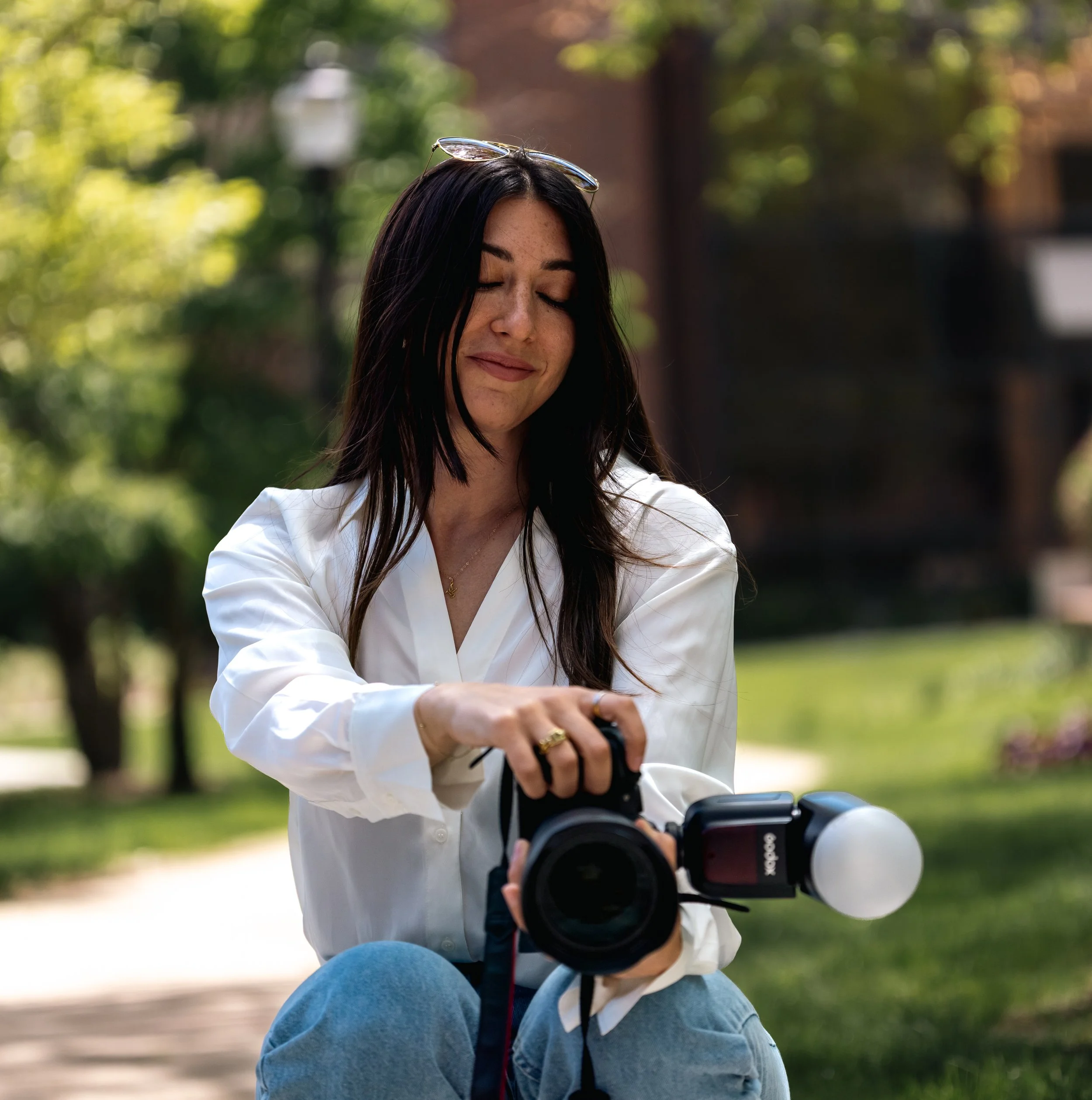 Young woman sitting outdoors in a park, with sunglasses resting on her head, holding a camera. She has long dark hair and is wearing a white shirt and blue jeans. The background is blurred with green trees and sunlight.