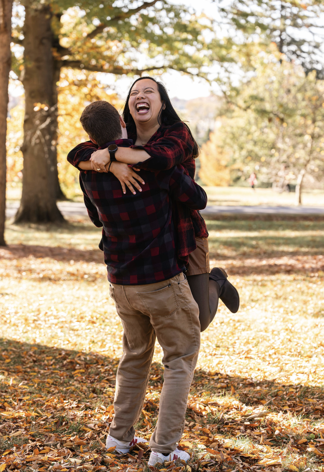 A couple is hugging and dancing in a park during autumn, surrounded by fallen leaves, with trees in the background.