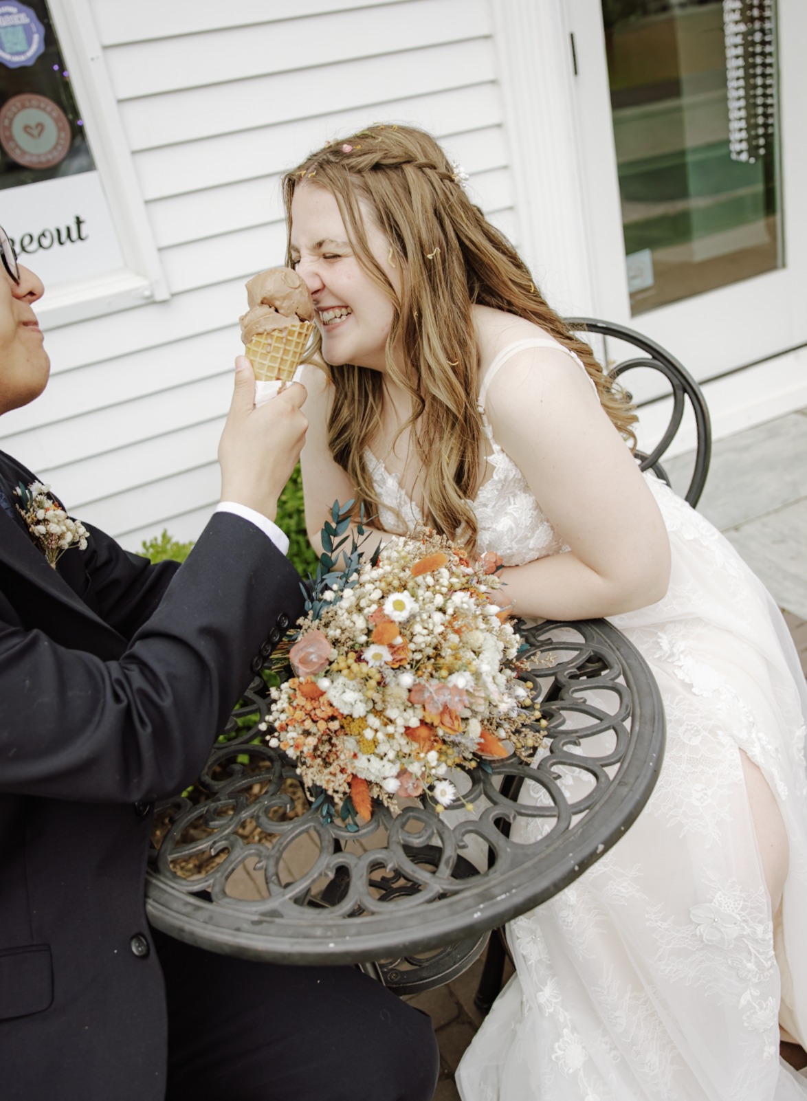 A bride and groom share a playful moment outdoors, with the groom holding an ice cream cone near the bride's face as she smiles and nuzzles it, sitting at a decorative metal table with a colorful bouquet of flowers.