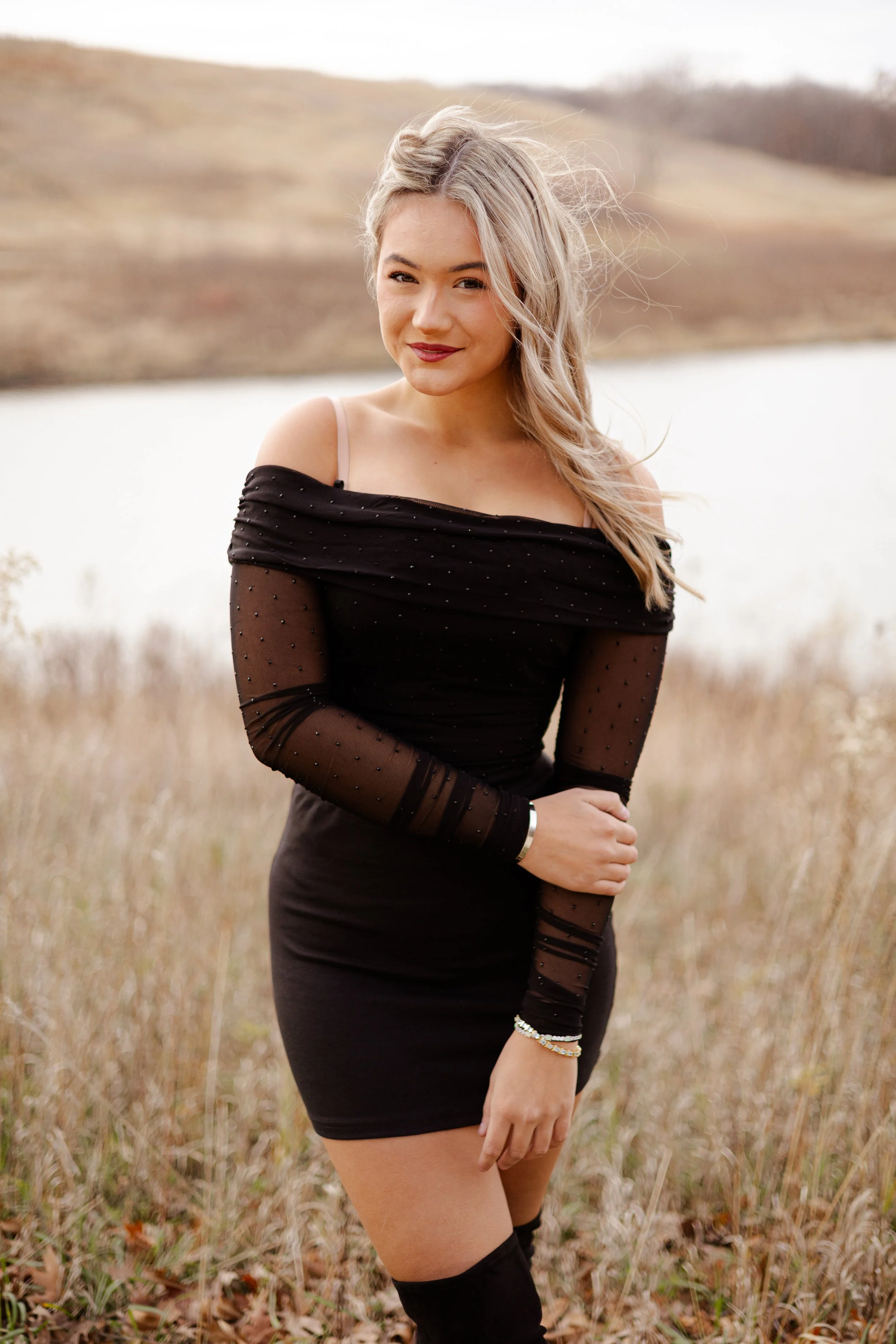 A young woman with blonde hair and light skin standing outdoors in a grassy area near a lake or river, wearing a black off-the-shoulder dress with sheer, polka-dotted long sleeves and black thigh-high stockings, looking at the camera with a slight sm