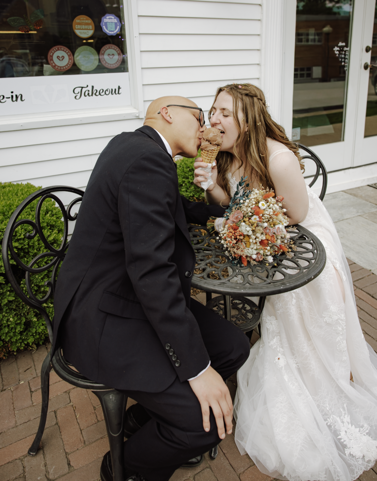 A wedding couple sitting at a table outside, sharing a chocolate ice cream cone. The bride is wearing a white wedding dress and holding a bouquet of flowers, while the groom is dressed in a black suit and glasses. There is a white building with a win