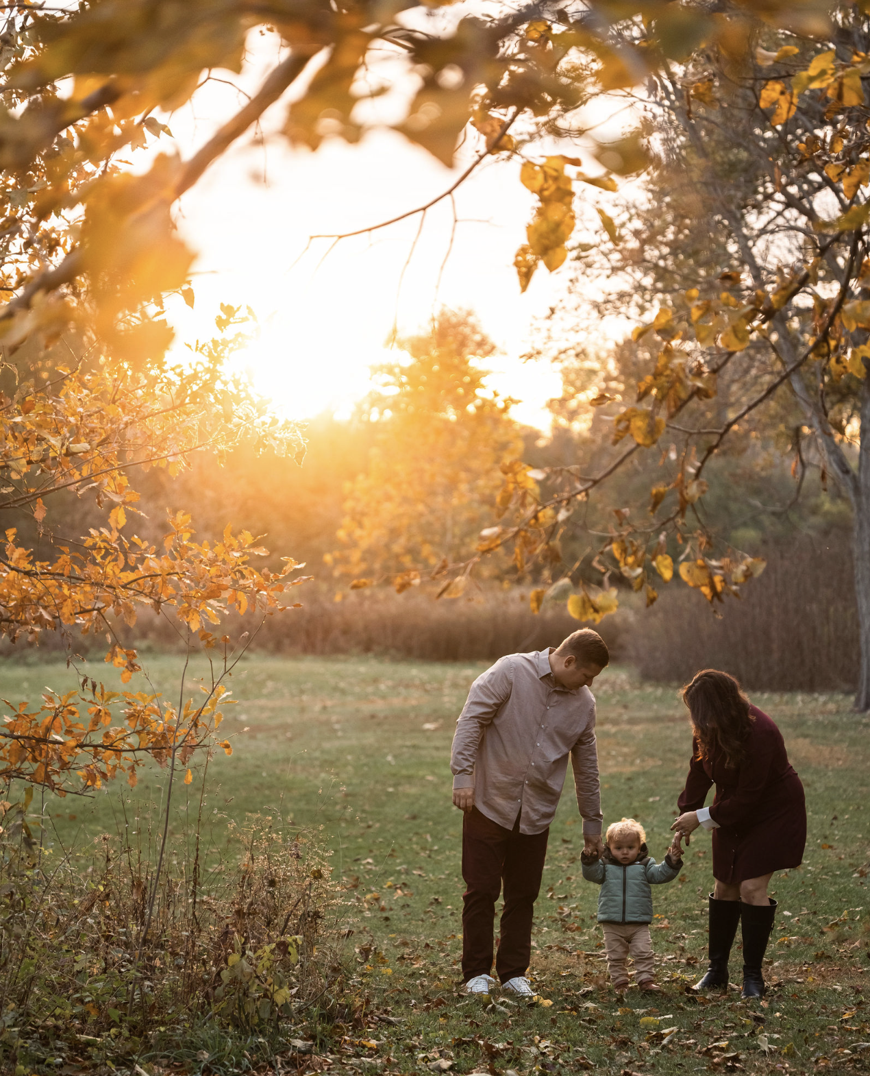 A family of three, a man, a woman, and a young child, enjoys a walk in a park during autumn at sunset. The man and woman hold the child's hands as they walk on grass covered with fallen leaves, with trees and orange leaves in the background.