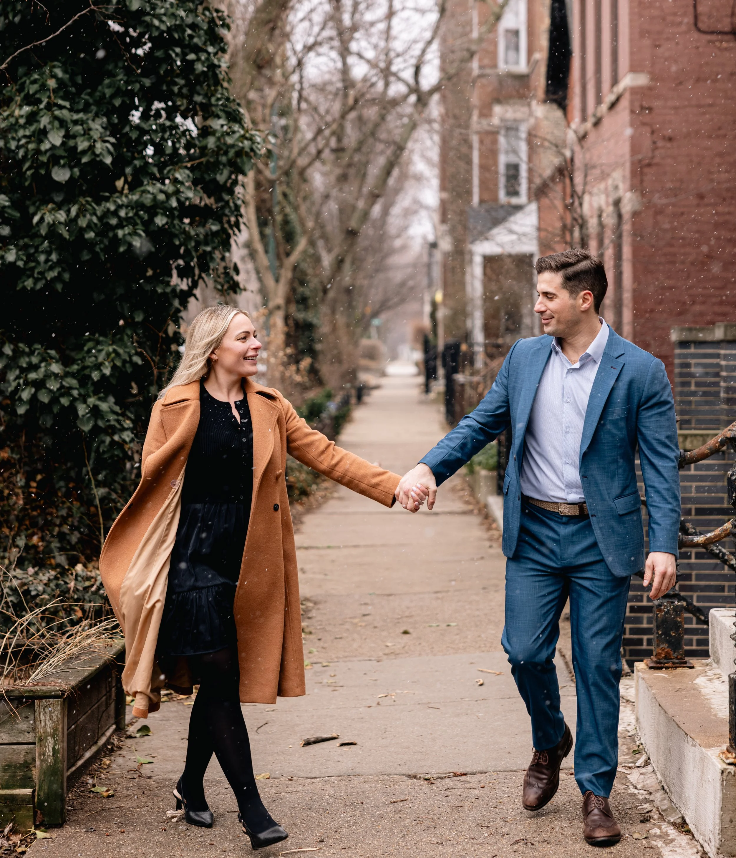 A man and woman holding hands walking on a sidewalk in a city neighborhood, smiling at each other, with trees, houses, and falling snow in the background.