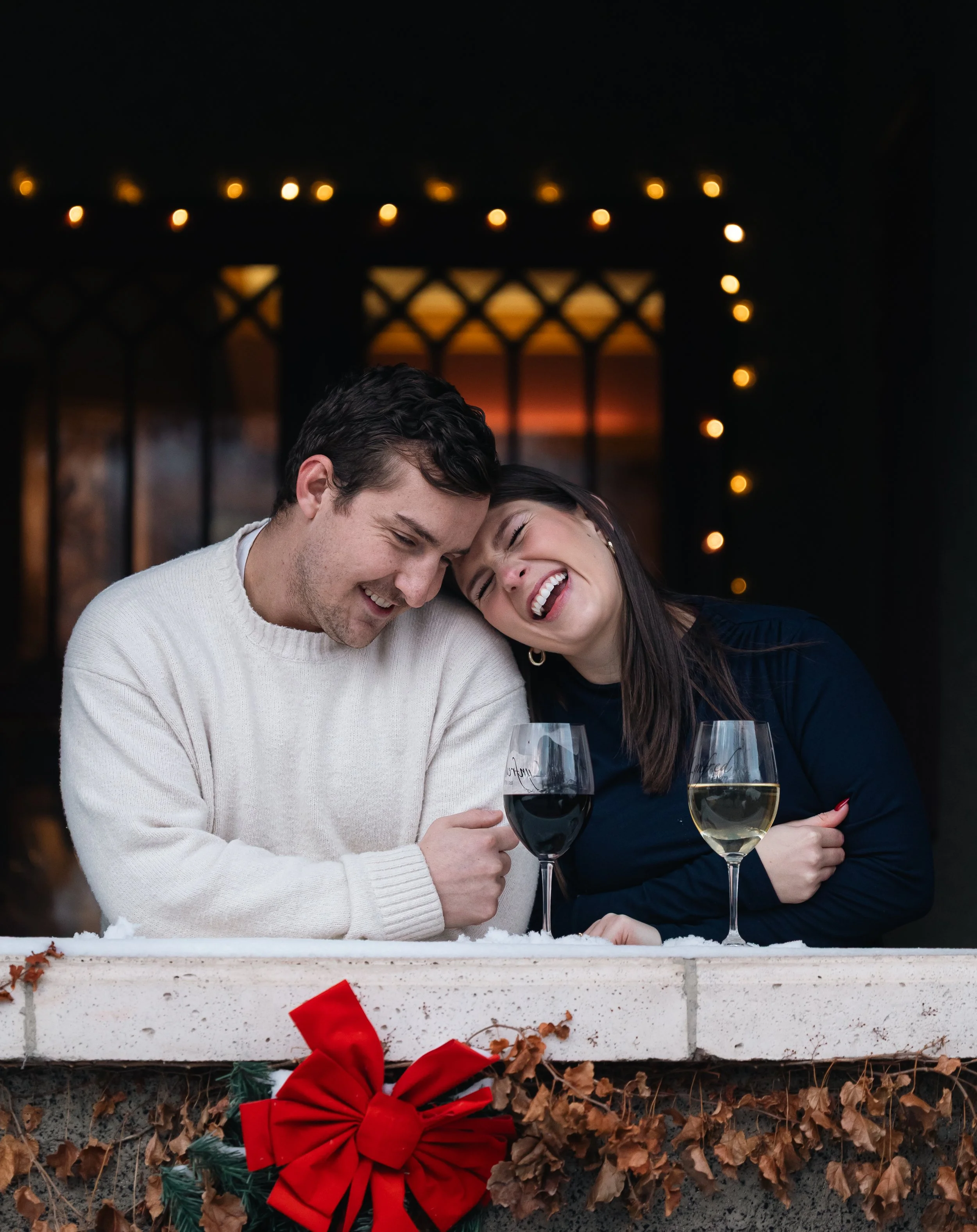 A couple smiling and leaning their heads together, each holding a glass of white wine, with a Christmas wreath and fallen leaves outside.