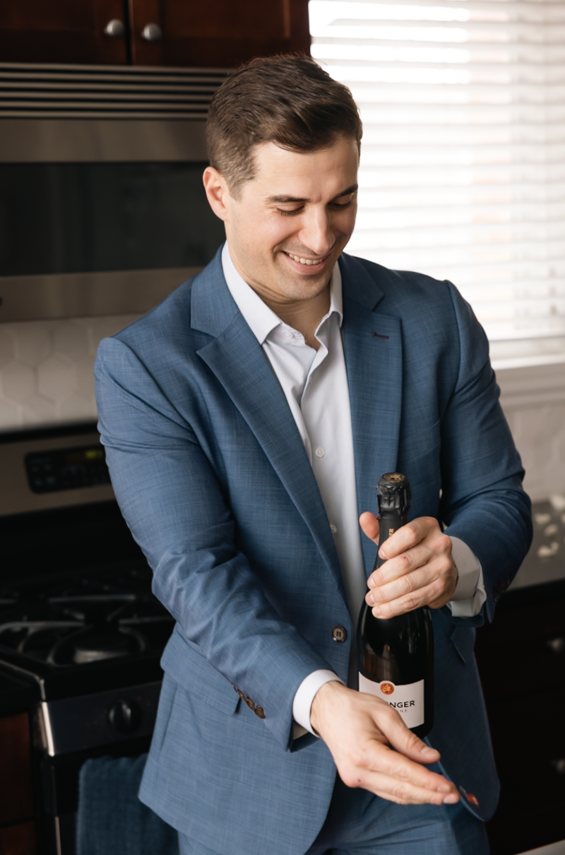 A man in a blue suit opening a bottle of champagne and smiling.