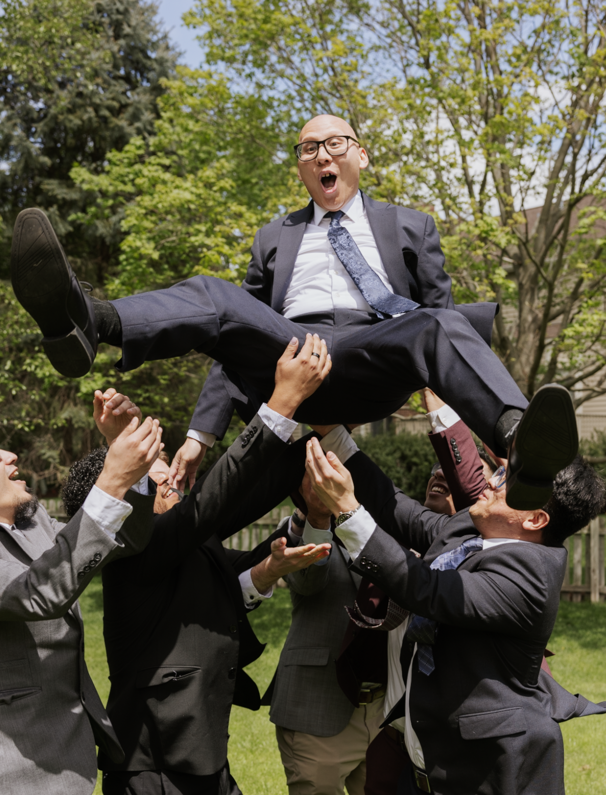 Group of people in business suits forming a human pyramid outdoors, lifting a man wearing glasses and smiling, against a background of trees and blue sky.
