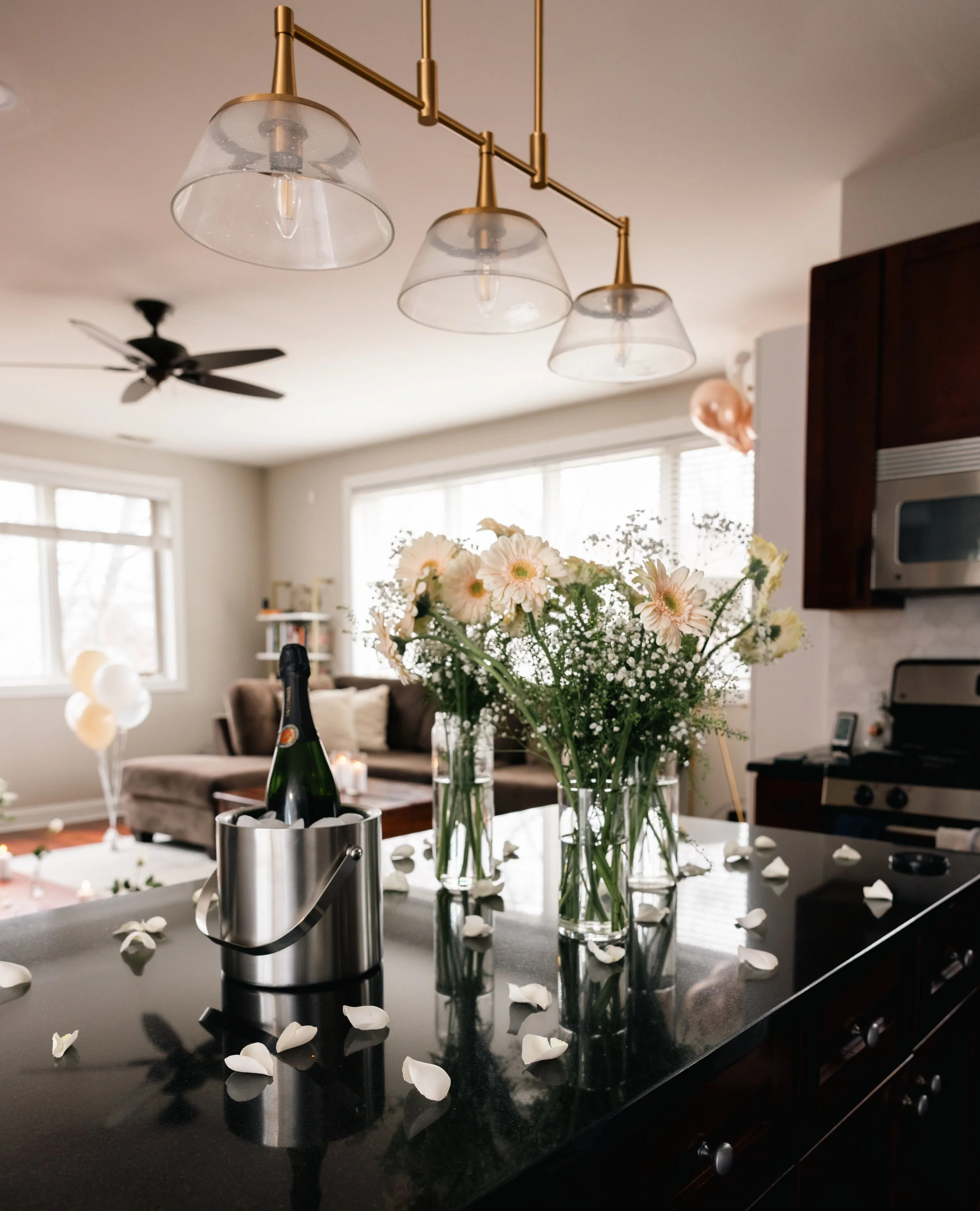 A modern kitchen with a black countertop, clear glass flower vases with white and pale pink flowers, scattered rose petals, a metallic champagne bucket with a bottle inside, and a lighting fixture with three glass shades hanging from a brass rail. In