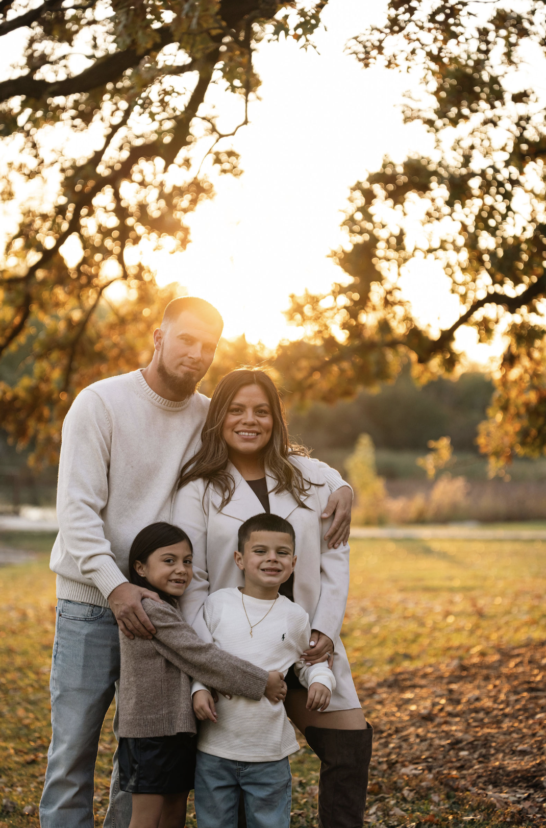 A diverse family of five smiling outdoors during sunset, surrounded by autumn trees and fallen leaves.