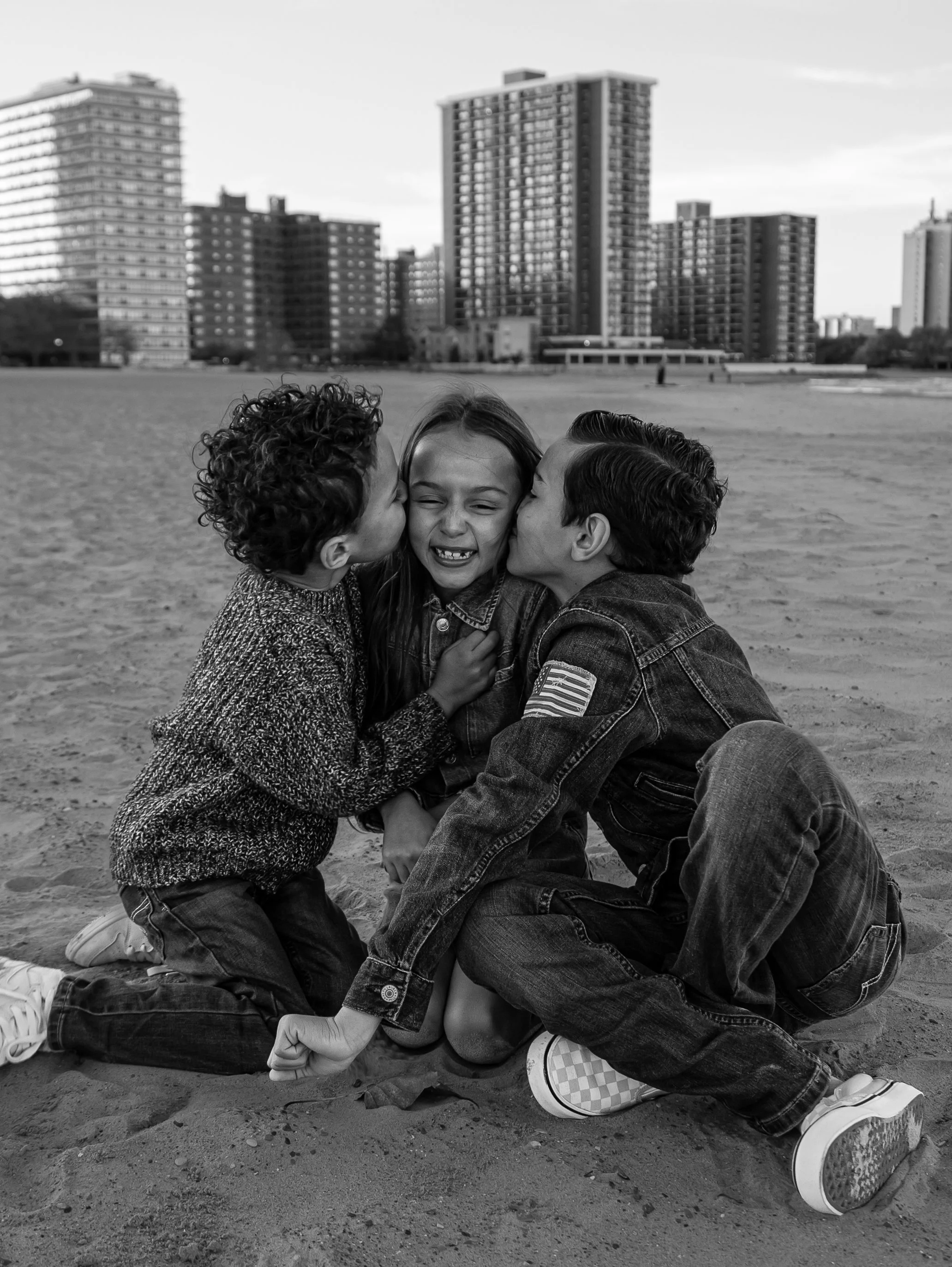 Three children sitting on a sandy surface, with two boys giving a kiss on the cheeks to the girl in the middle. The scene is set outdoors in front of a background of tall buildings.