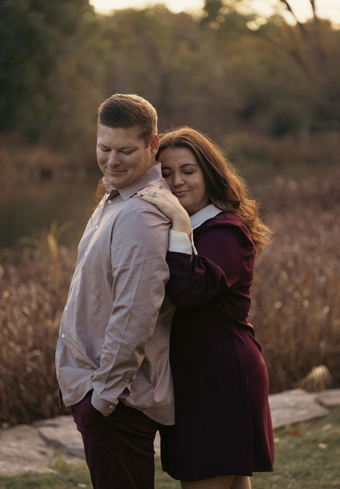 A young couple hugging outdoors at sunset, with the woman resting her head on the man's shoulder and both showing peaceful expressions.