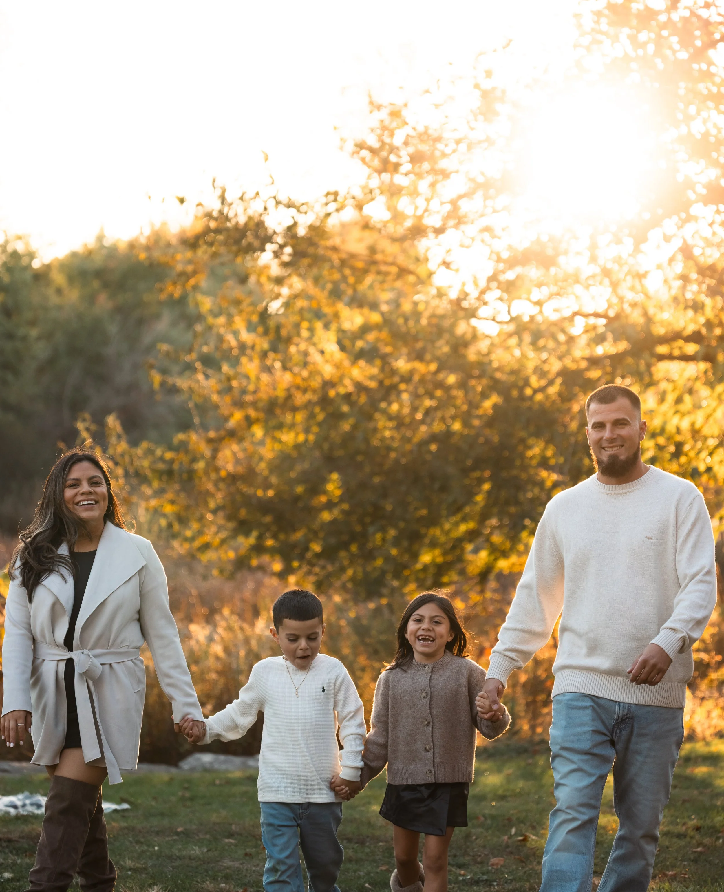 A group of people walking outdoors during sunset, holding hands, with smiling faces. The group includes a woman with long hair, two young children, and a man, all dressed casually. The background features trees and warm sunlight.
