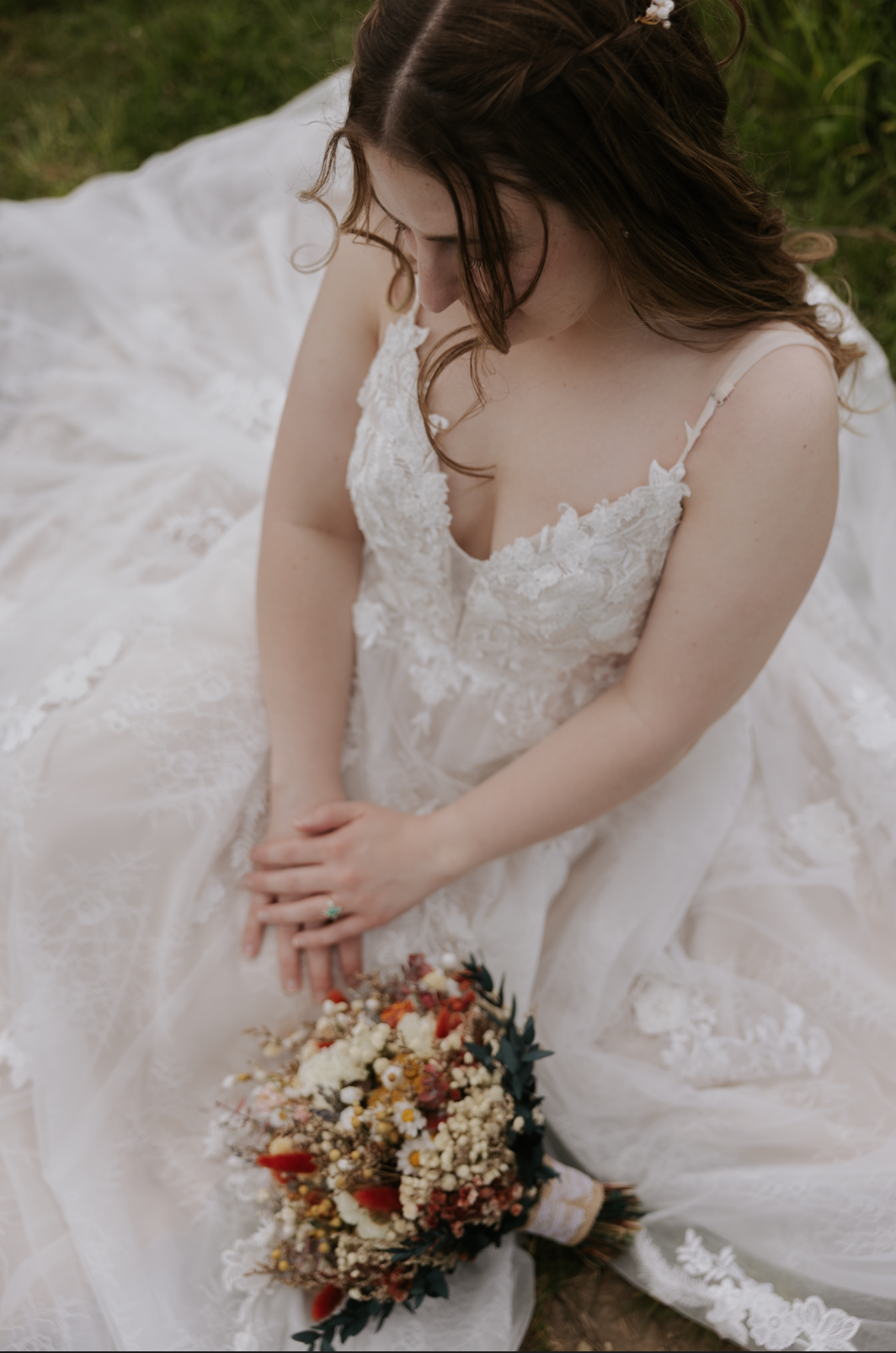 A woman wearing a lace wedding dress sitting outdoors on the grass, holding a bouquet of flowers.