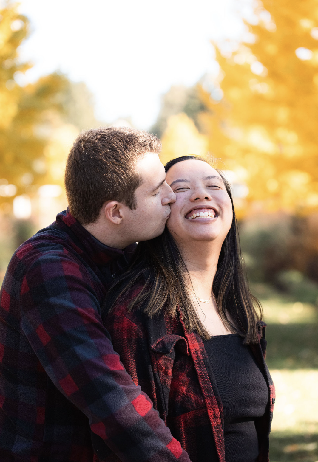 A young man kissing a young woman on the cheek outdoors during autumn, both smiling with yellow trees in the background.