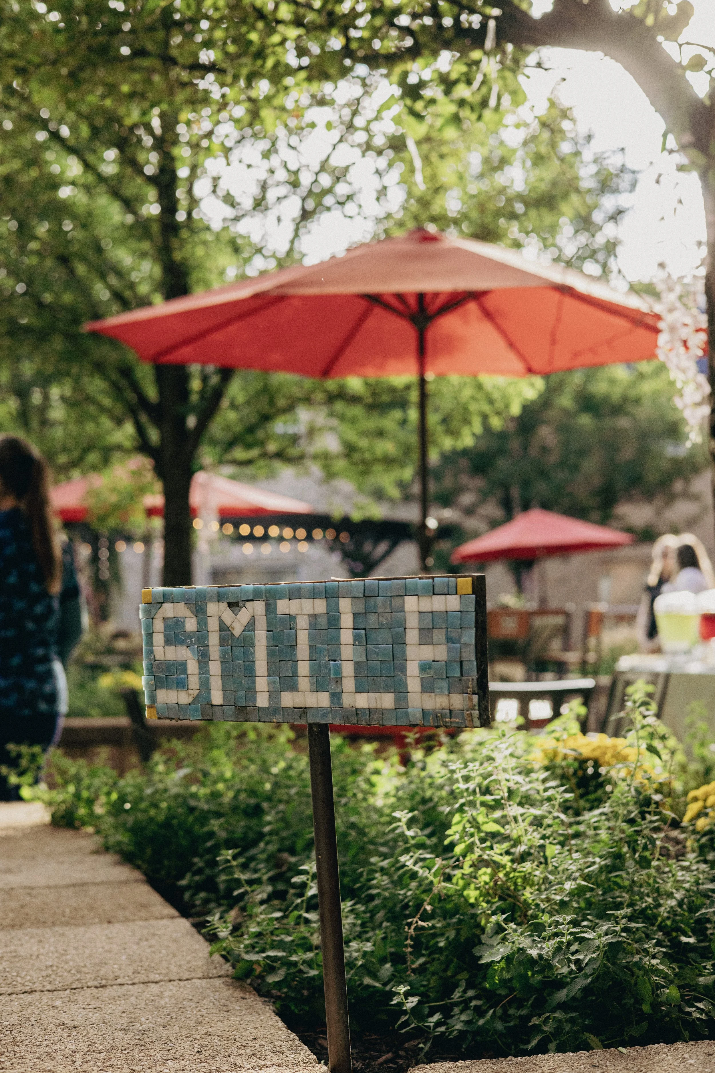 A mosaic sign on a metal post with the word "GARDEN" on it, set in a lush outdoor setting with red umbrellas and people in the background.