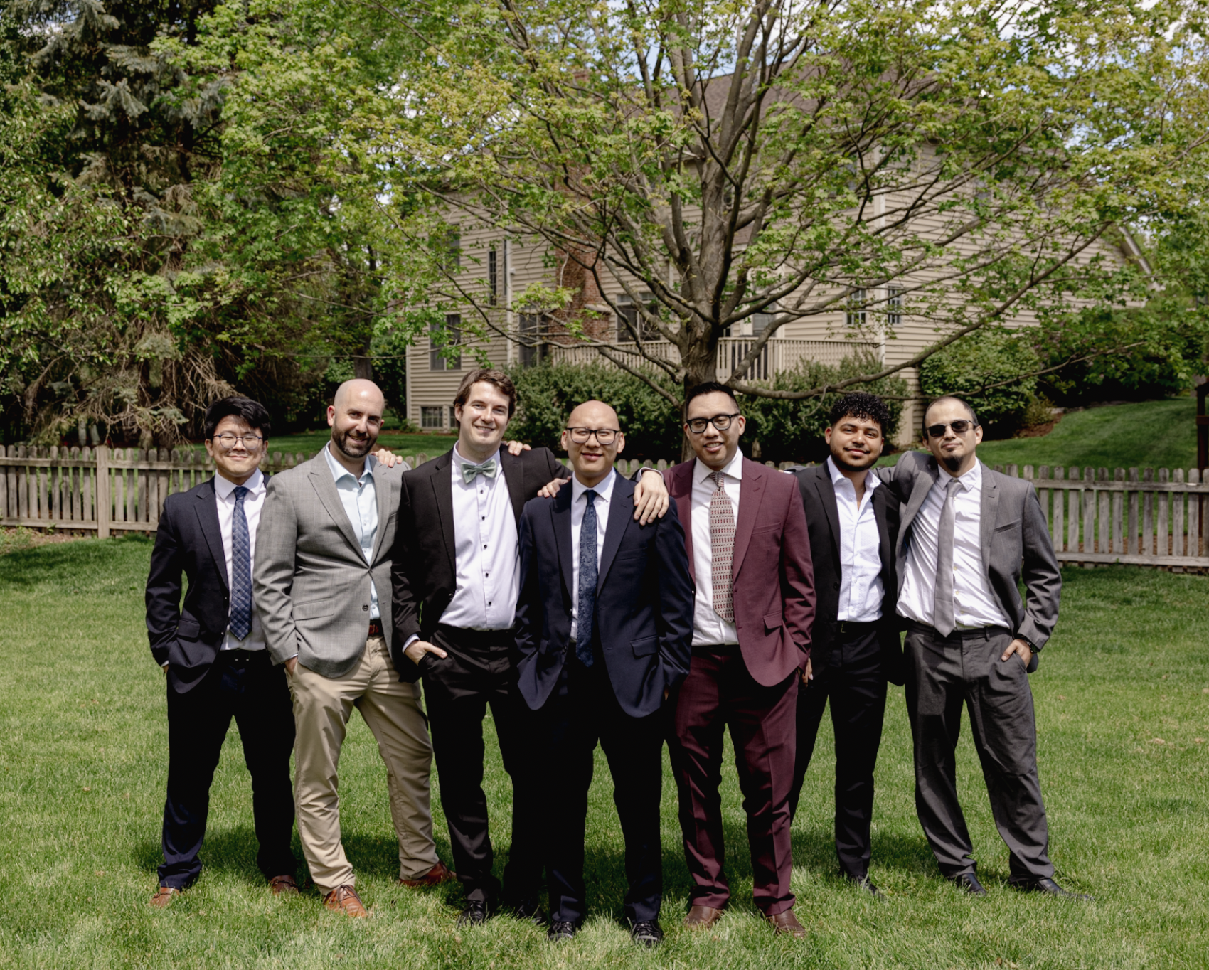 A group of seven men dressed in suits and ties posing outdoors on a grassy lawn with trees and a house in the background.