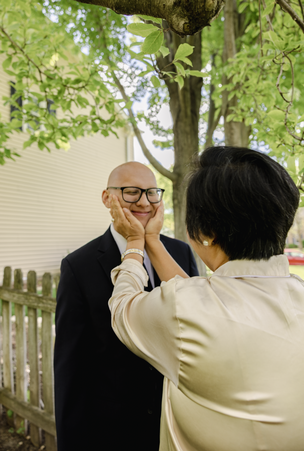 A woman gently holds a man's face, making him smile with his eyes closed outdoors under a large leafy tree, with a wooden fence and house in the background.