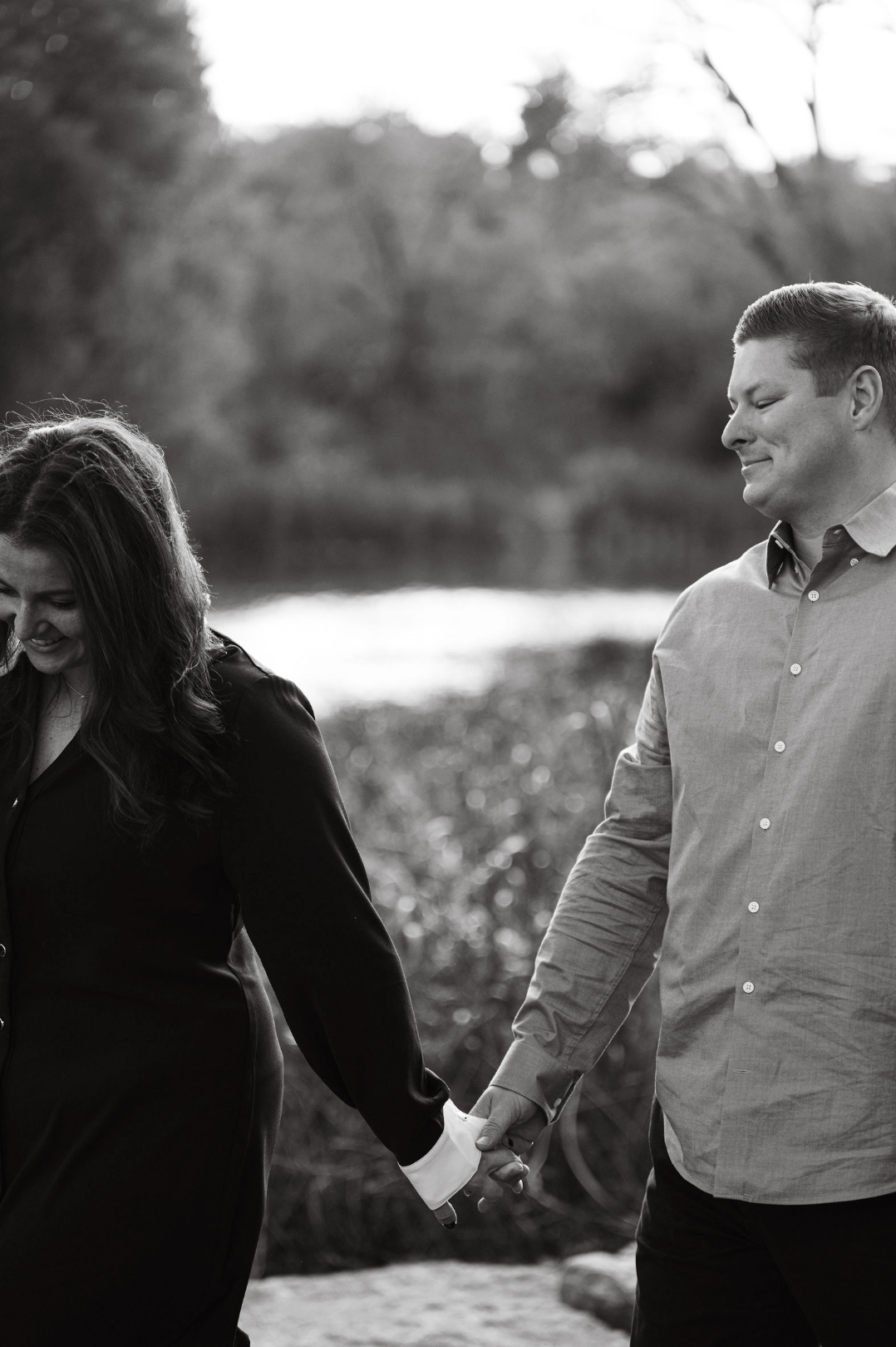 A black and white photo of a man and woman holding hands outdoors, both smiling and looking at each other, with a blurred natural background.
