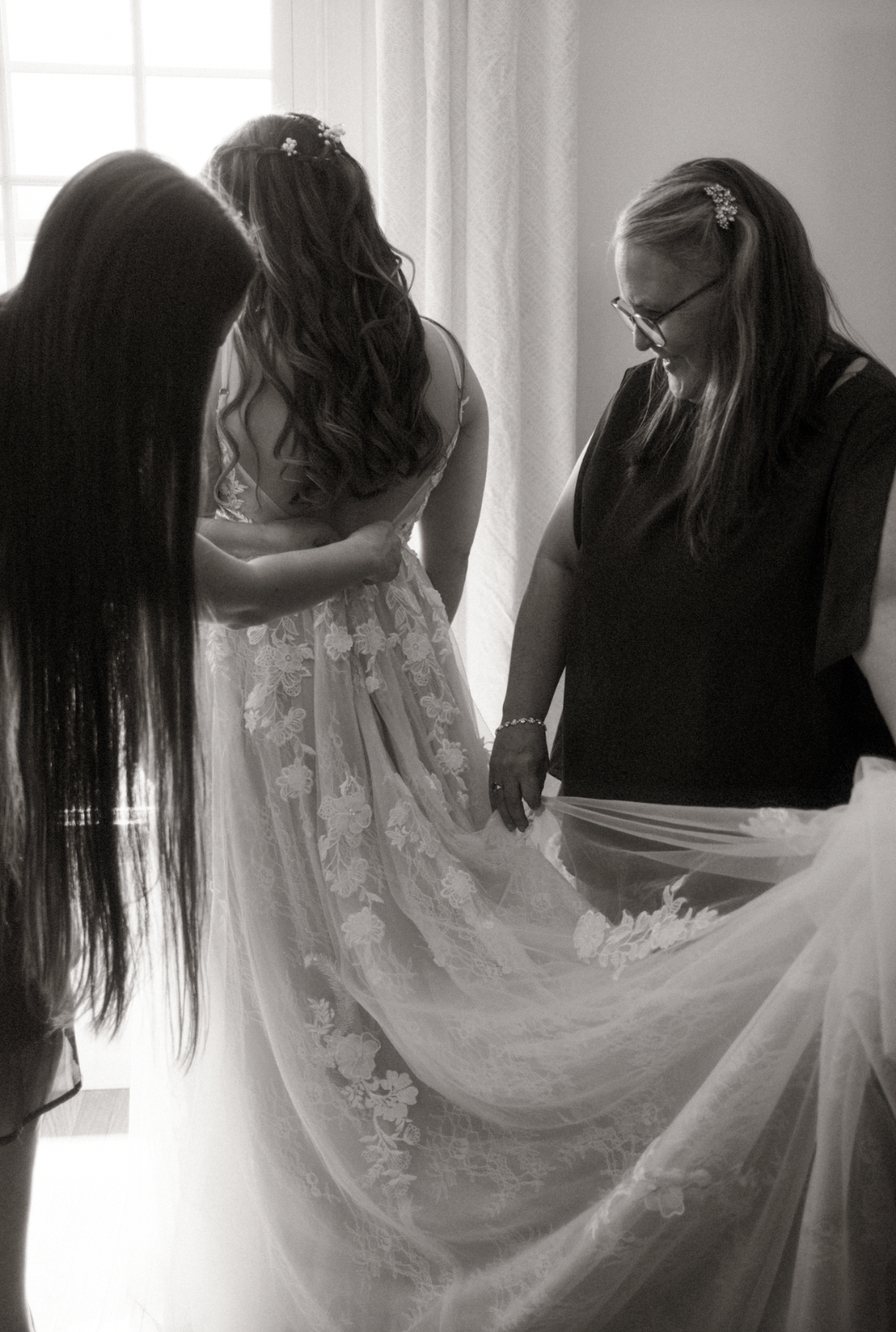 Women helping a bride get ready, adjusting her wedding dress, in a well-lit room.