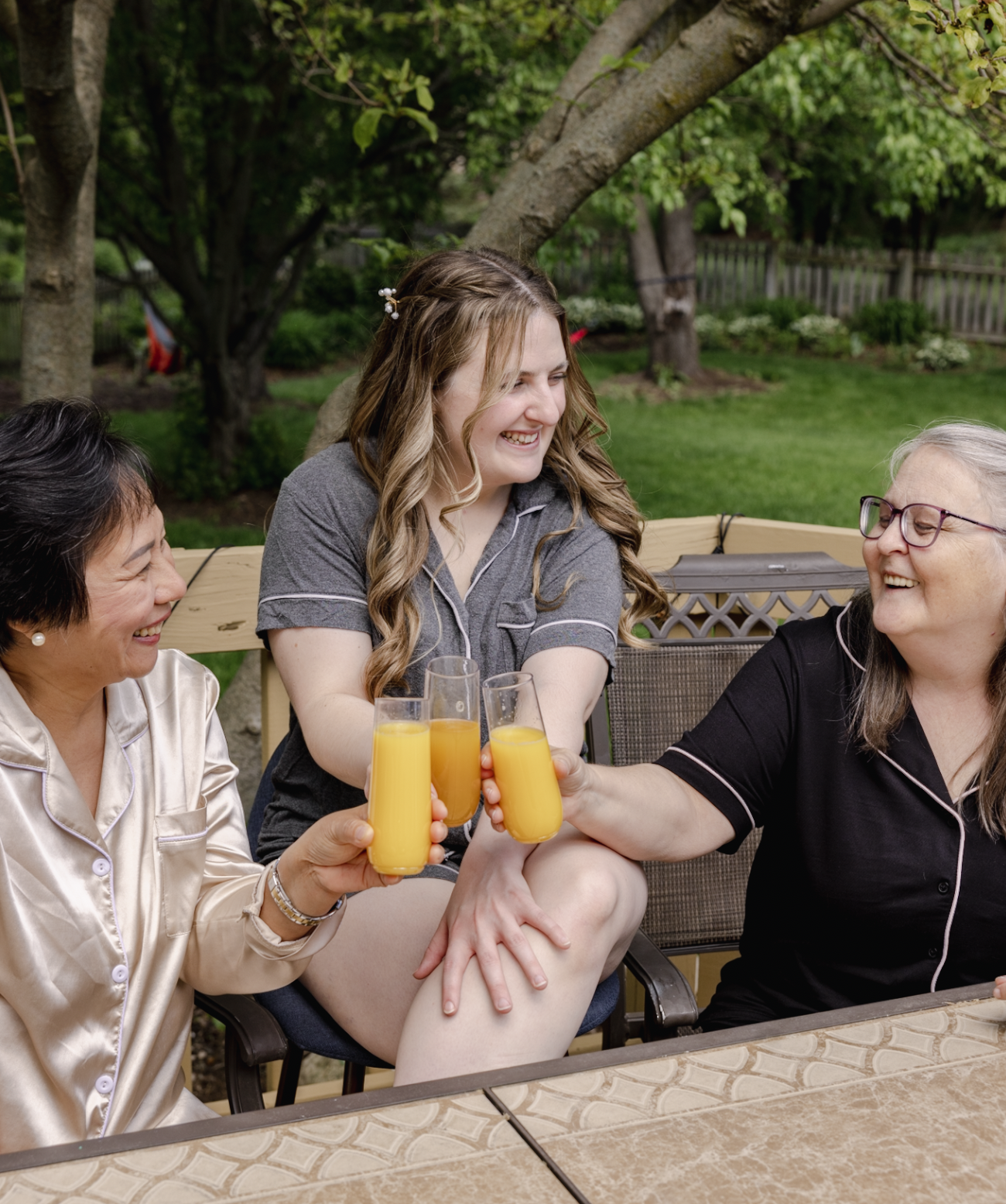 Four women sitting outdoors, smiling and clinking glasses of orange juice, enjoying a sunny day in a garden.