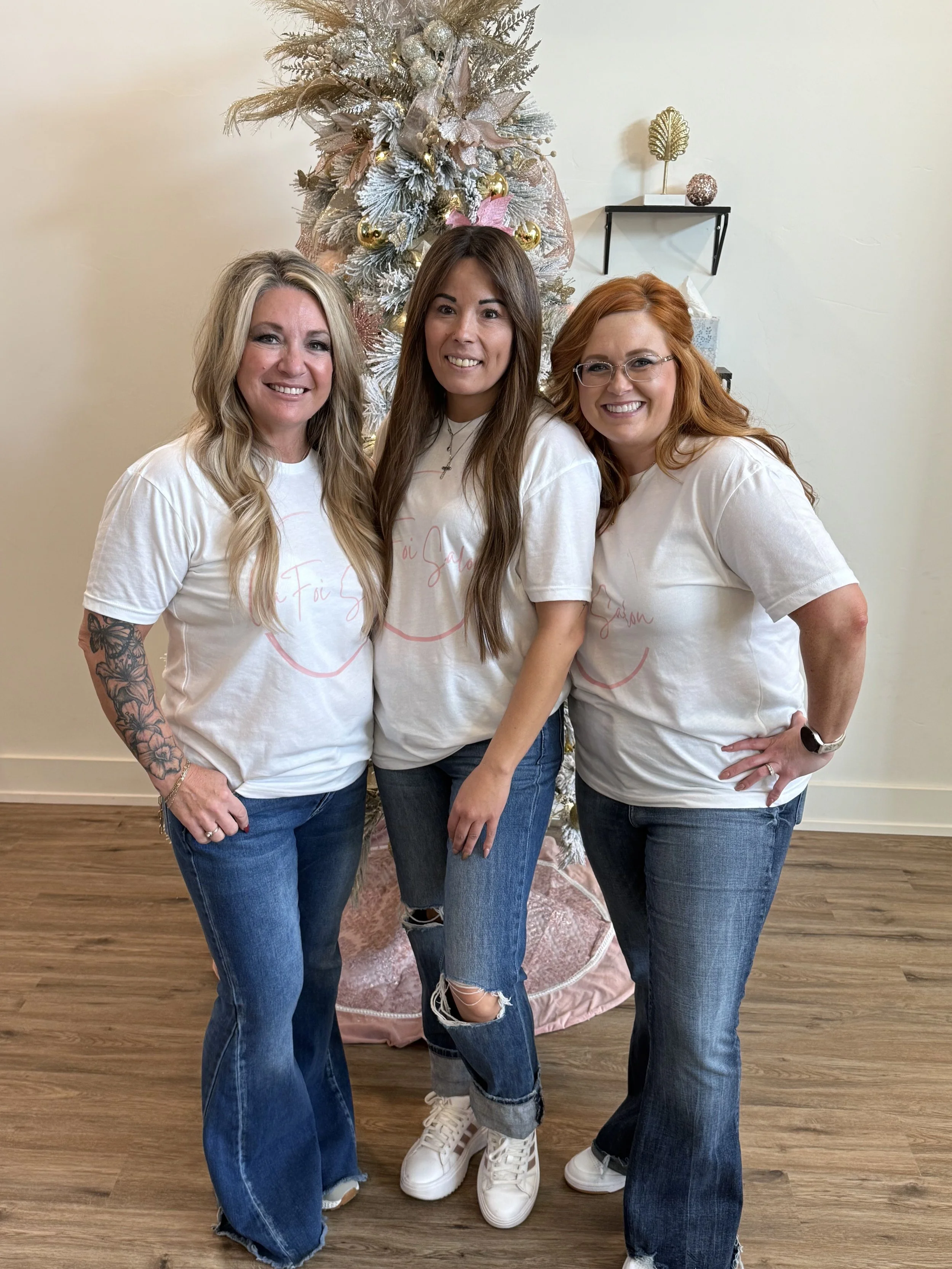 Three women standing in front of a decorated Christmas tree, smiling, wearing matching white t-shirts and jeans.