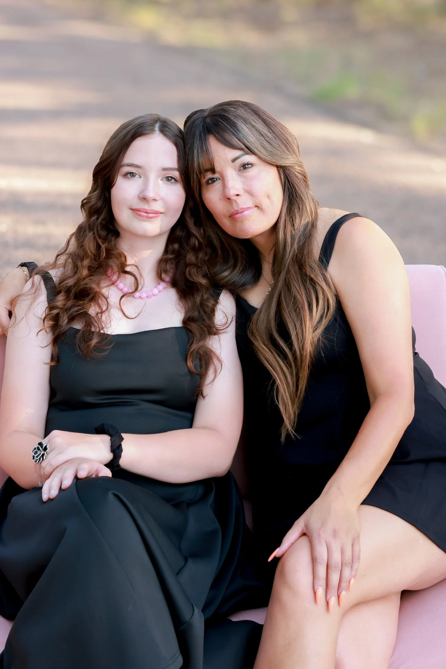 Two women sitting closely together outdoors, with one woman resting her hand on the other's leg. They are wearing black dresses and smiling softly at the camera, with a blurred natural background.