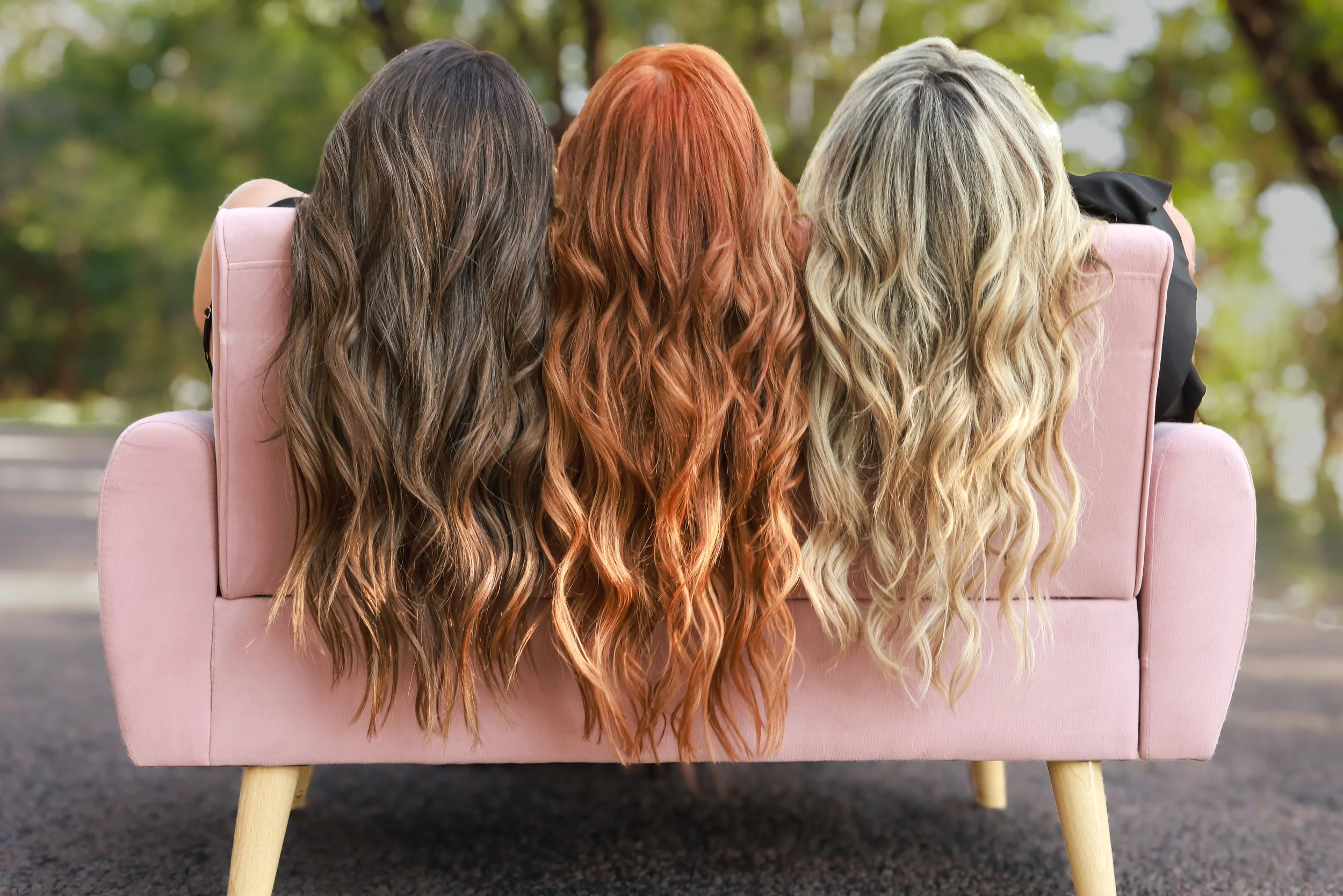 Three women with long wavy hair, viewed from behind, sitting on a pink sofa outdoors in a park with green trees.