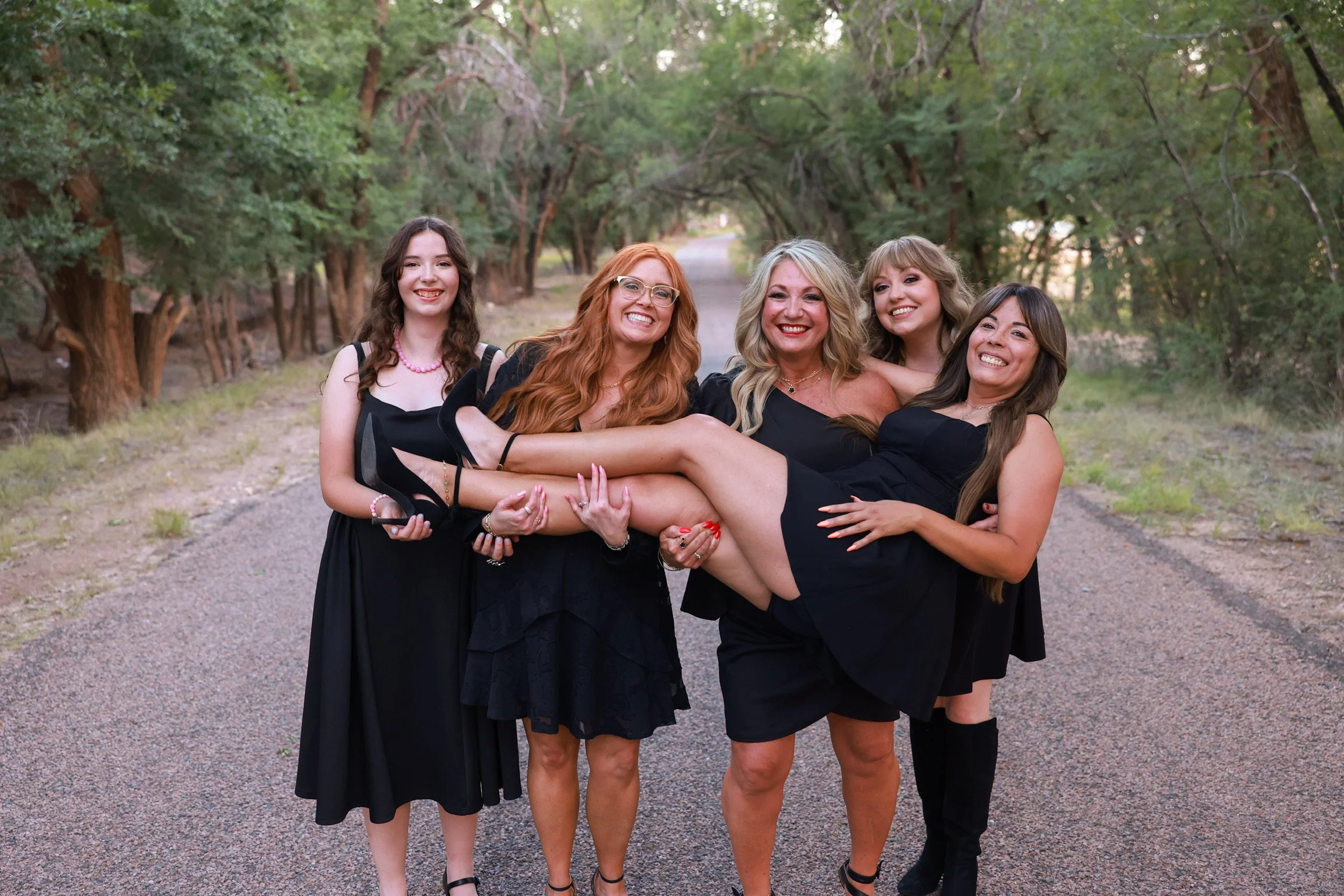 A group of five women dressed in black, smiling and standing together on a gravel path in a wooded area, with trees and greenery around them.