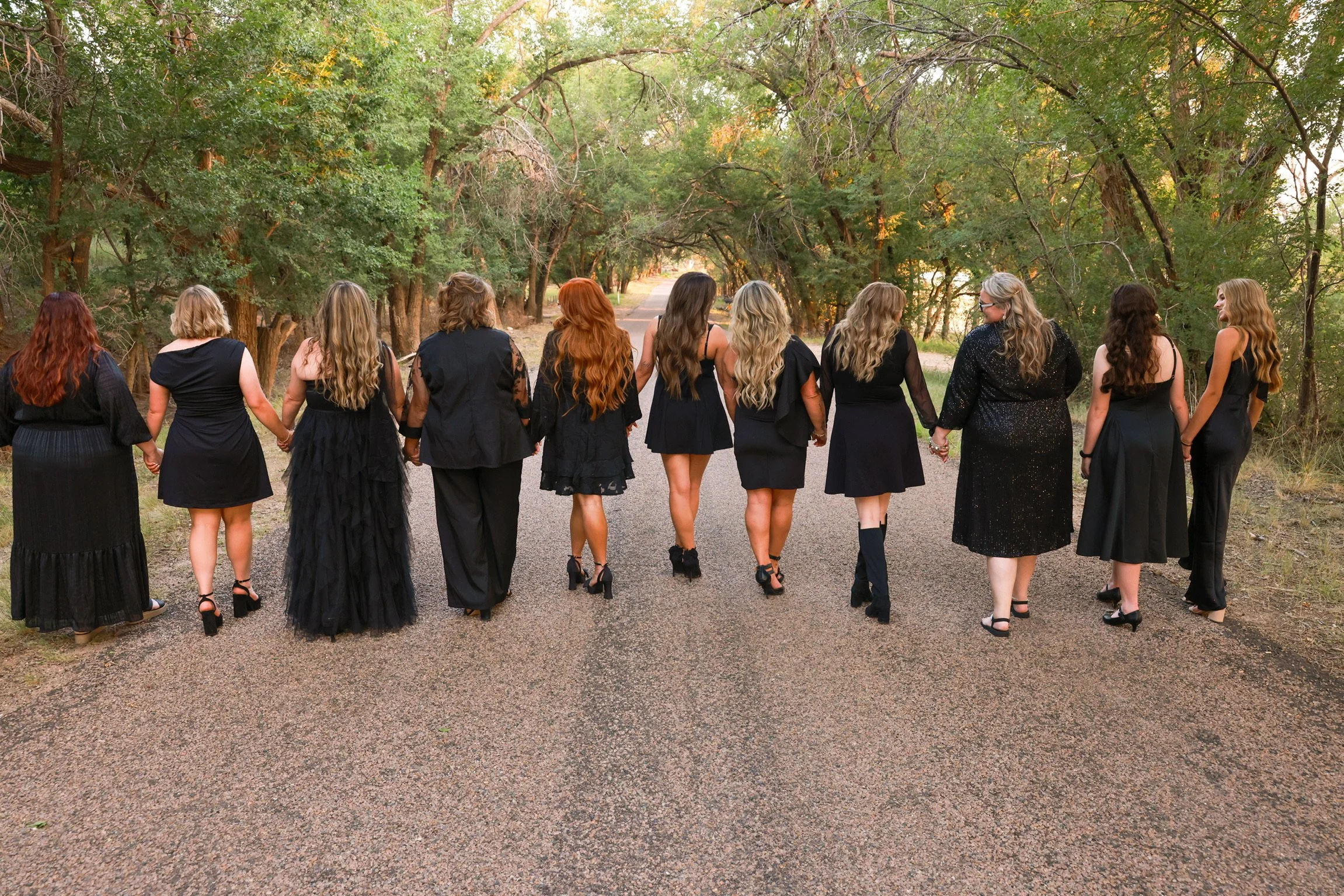 A group of women dressed in black, holding hands and walking down a gravel path surrounded by green trees.